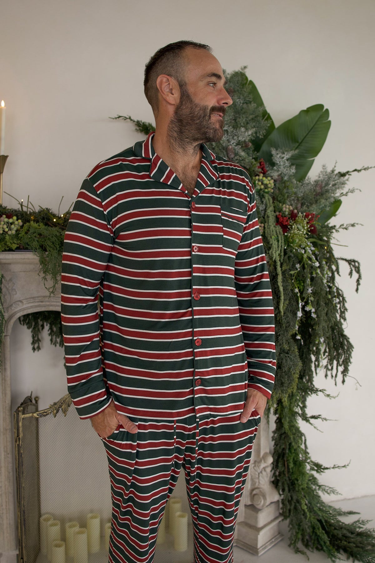 A man wearing a forever french baby Men’s Bamboo Button Up Lounge Set in Holiday Stripe, featuring a long-sleeved button-up shirt and matching pants with a red, green, and white horizontal stripe pattern.