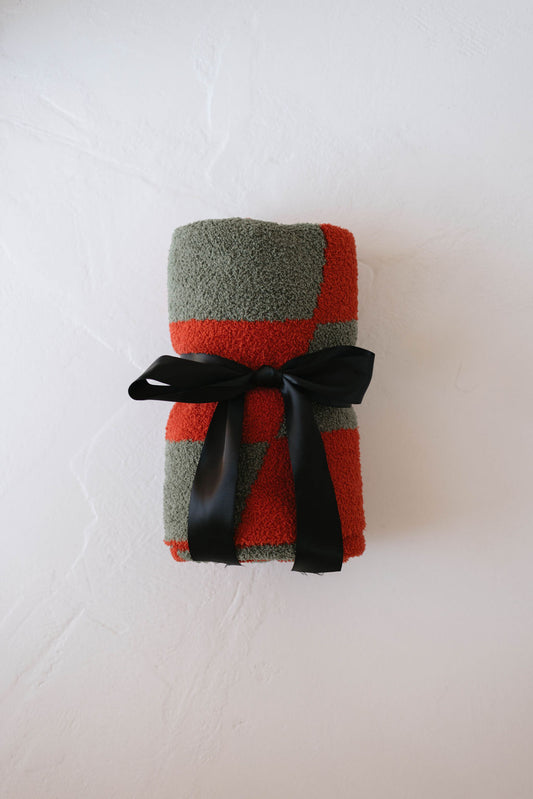 A folded forever french baby Retro Christmas Plush Blanket featuring a red and gray checker pattern is elegantly tied with a black ribbon, resting on a white textured surface.