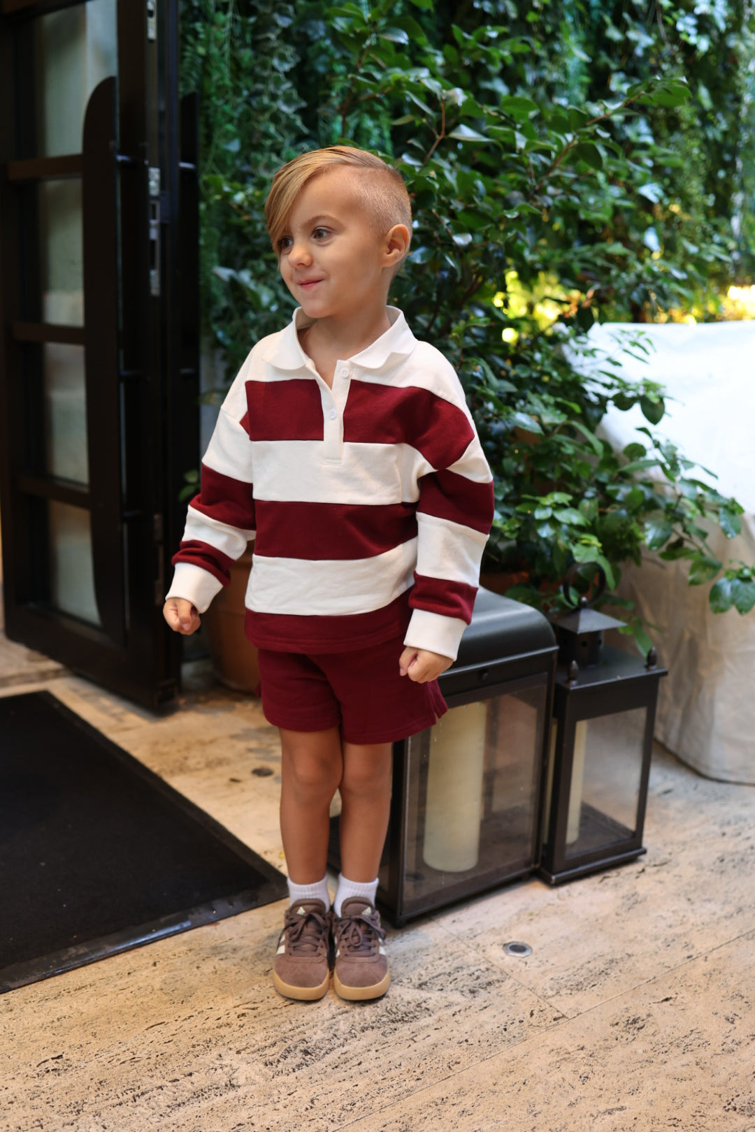 A young boy wearing a white and maroon striped long-sleeved polo shirt and Child Terry Shorts | Deep Cranberry.