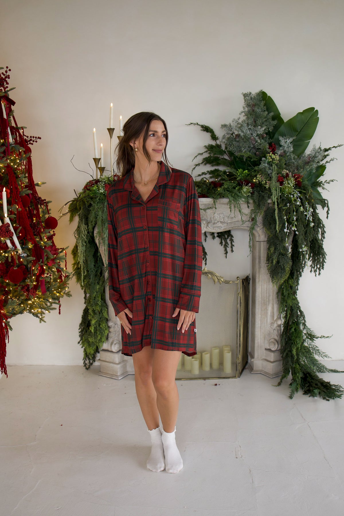 A woman in a Women’s Bamboo Sleeping Dress | Christmas Plaid and white socks stands in front of a fireplace decorated for the holidays.