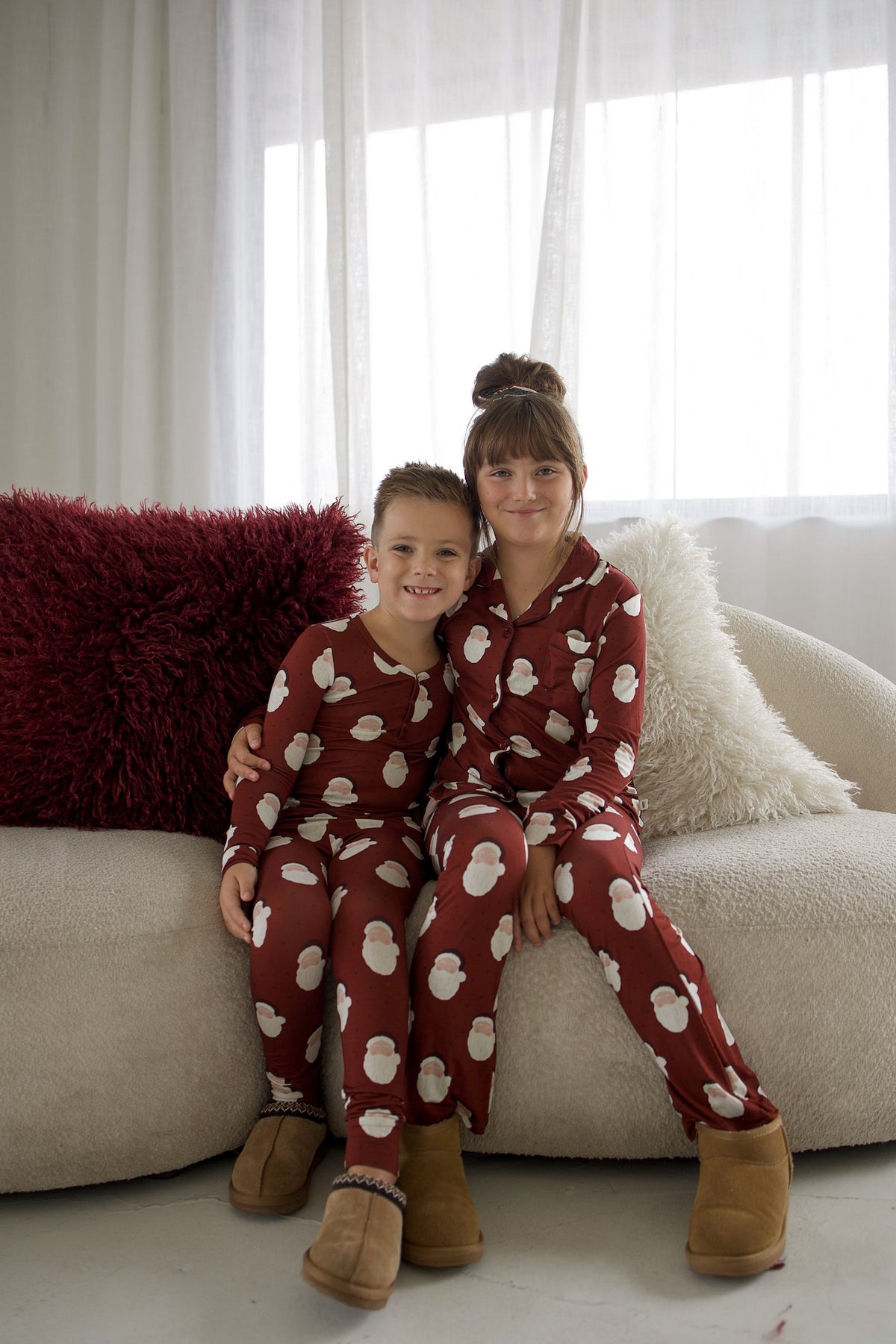 A boy and a girl wearing matching Girl’s Bamboo Button Up Lounge Set | Vintage Santa burgundy sets with a Santa Claus print, sitting together on a sofa.