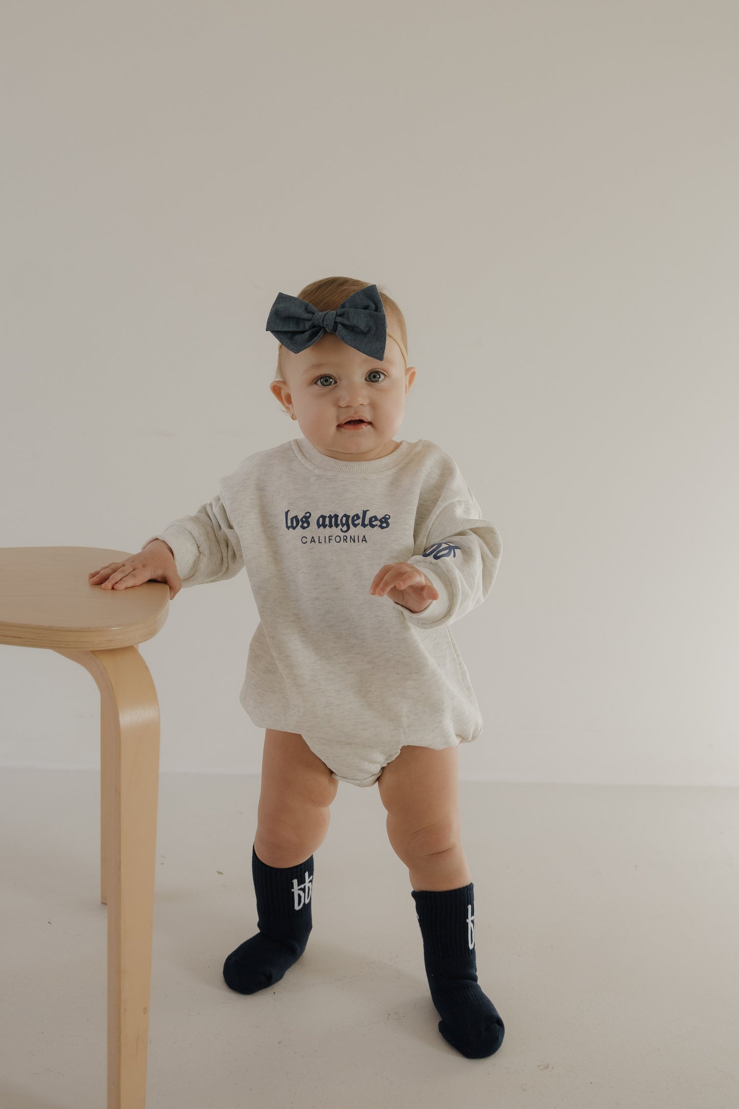 A baby wearing the forever french baby "Sweatshirt Romper | ff Los Angeles" in a light color, black knee-high socks, and a large dark bow headband stands by a light wooden table against a plain white background.