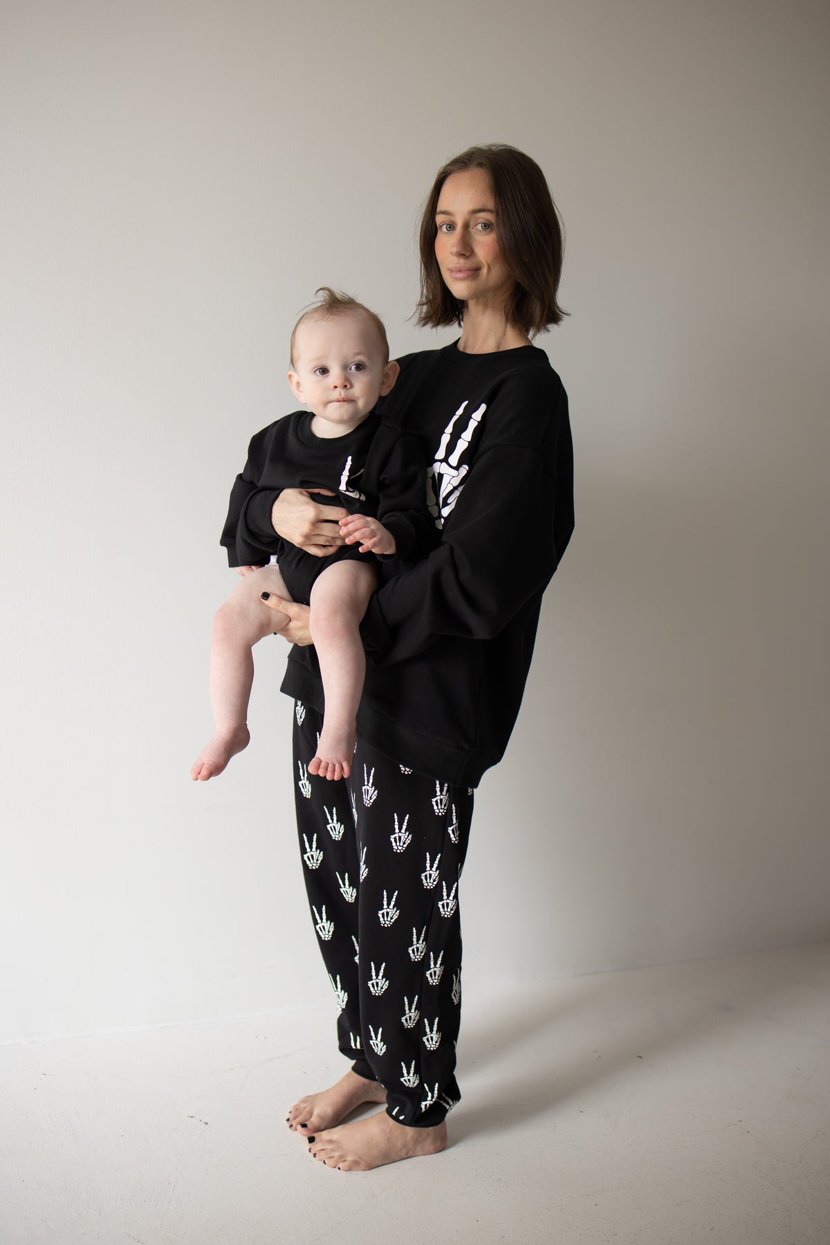 A woman stands barefoot holding a baby, both dressed in the forever french baby Women’s Sweat Set | Midnight Skelly—matching black sweatsuits adorned with white handprint designs—posing against a plain light-colored wall. The cozy sets make an ideal pumpkin patch outfit for this spooky season.