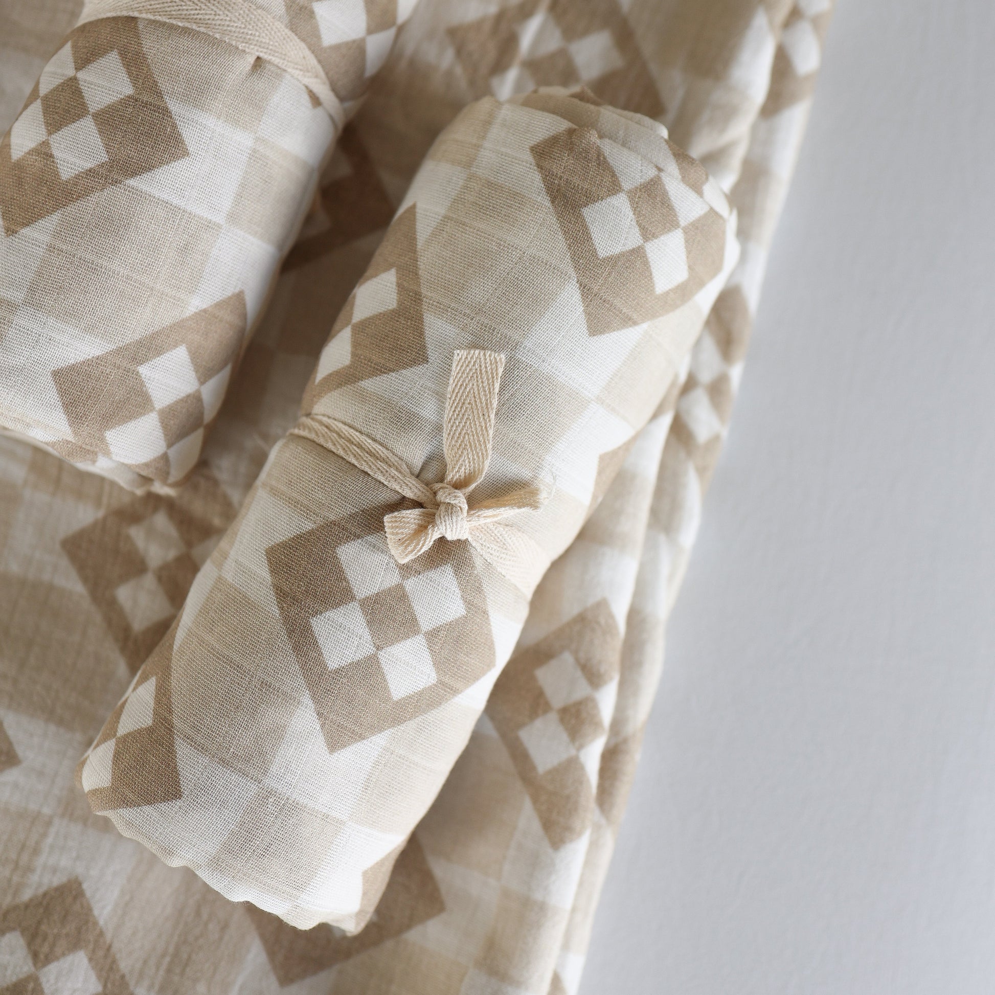 The Muslin Swaddle | Cottage Quilt by forever french baby features rolled beige and white geometric diamond-patterned muslin, tied with a matching fabric strip and placed atop a flat piece of the same material.