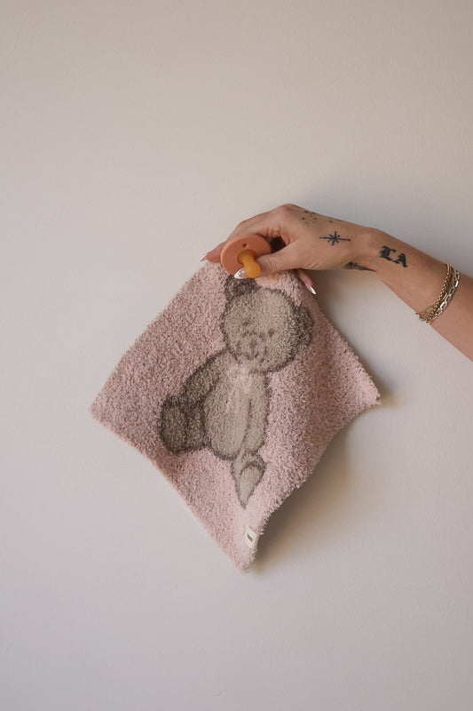 A tattooed hand wearing gold bracelets holds the Lovey | Lottie by forever french baby—a pink pacifier attached to a soft microfiber feather yarn blanket with a teddy bear design—against a plain, light-colored wall.