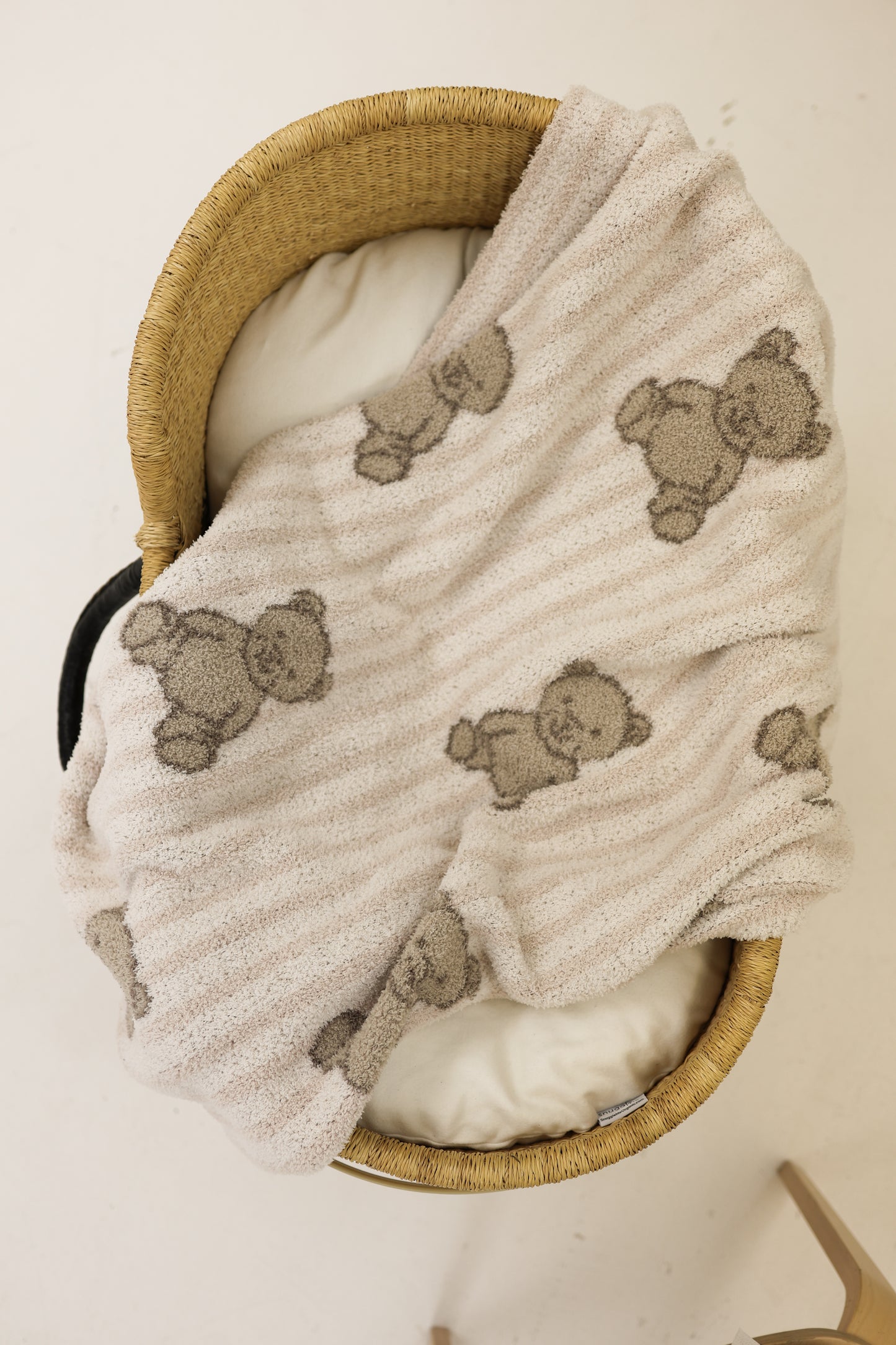 A woven bassinet with a white cushion is topped with the Plush Blanket | Teddy by forever french baby, an Oeko-Tex certified blanket featuring smiling teddy bears, all set against a light, neutral background.