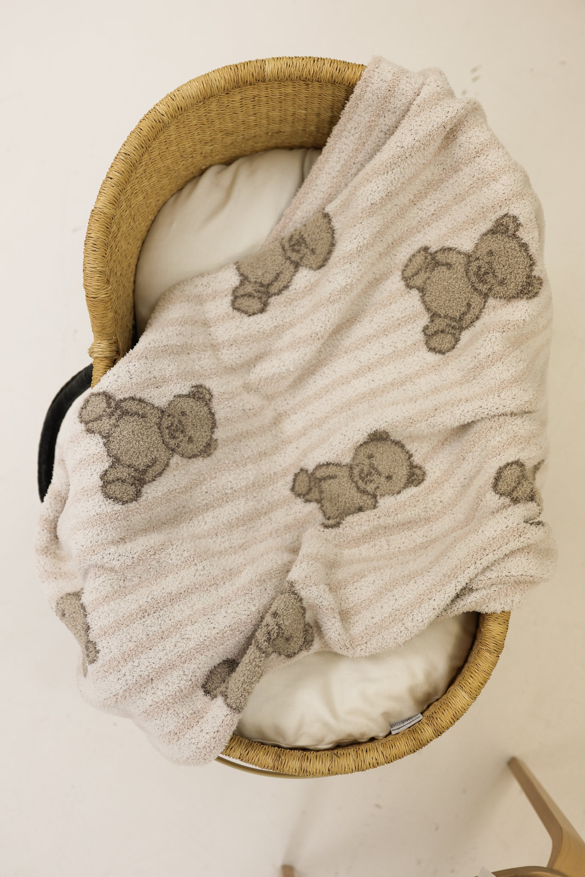 A woven bassinet with a white cushion is topped with the Plush Blanket | Teddy by forever french baby, an Oeko-Tex certified blanket featuring smiling teddy bears, all set against a light, neutral background.