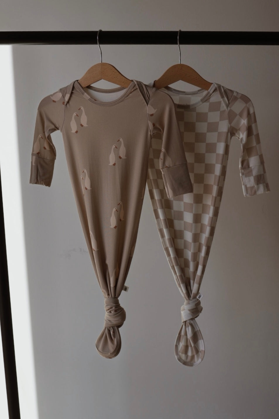 Two forever french baby Bamboo Knotted Gown | Silly Goose gowns in beige—one with a penguin print, the other checkered—hang on wooden hangers. Both hypoallergenic gowns are knotted at the bottom and displayed against a plain background.