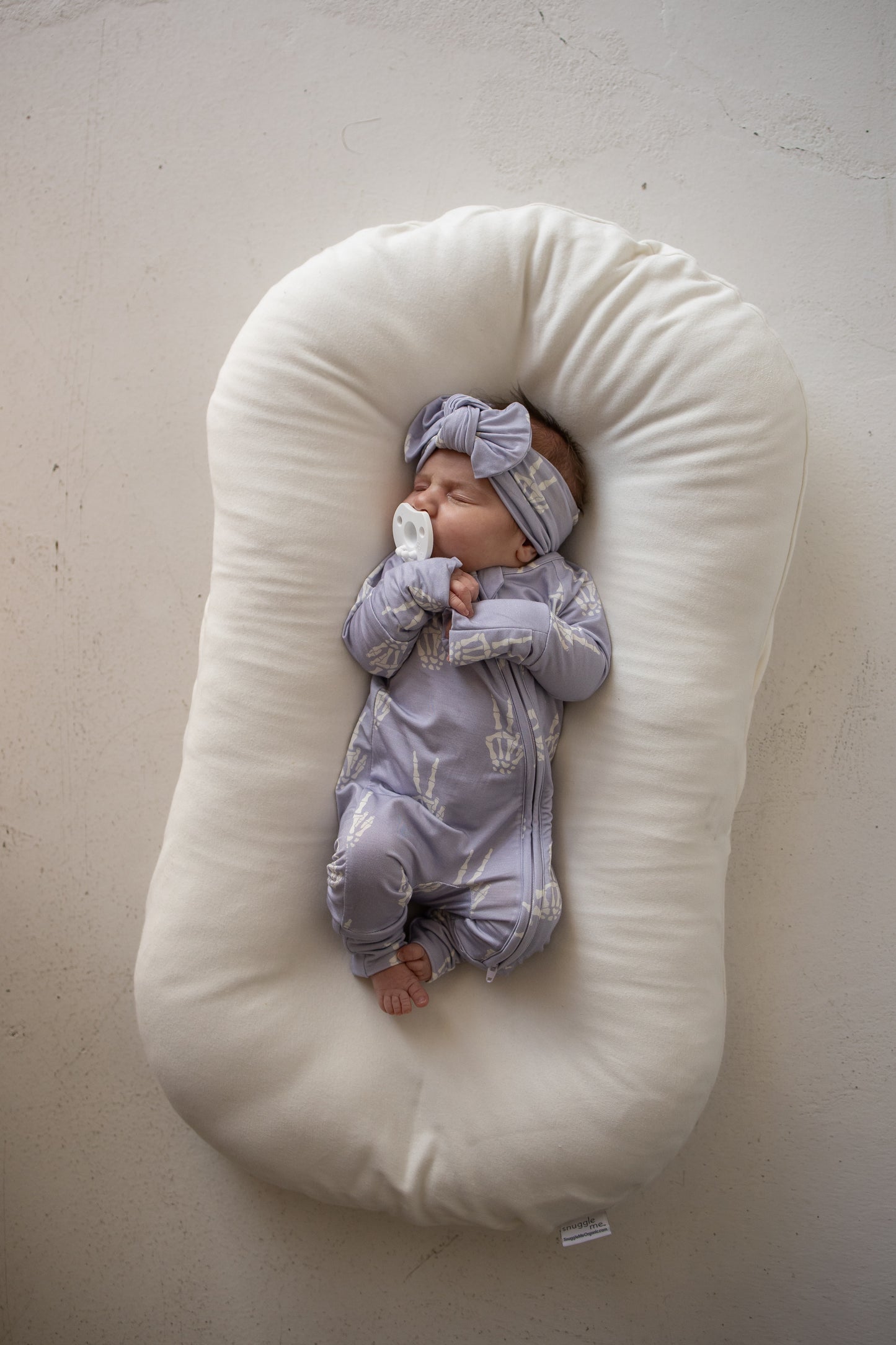A newborn baby wearing forever french baby’s Bamboo Zip Pajamas | Enchanted Skelly in soft, purple bamboo viscose and a matching headband sleeps peacefully on a white baby lounger while holding a pacifier.