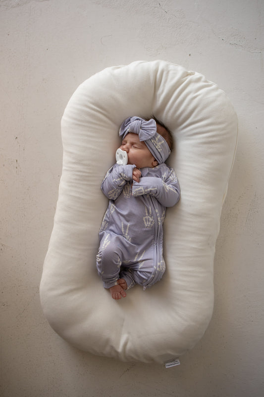 A newborn baby wearing forever french baby’s Bamboo Zip Pajamas | Enchanted Skelly in soft, purple bamboo viscose and a matching headband sleeps peacefully on a white baby lounger while holding a pacifier.