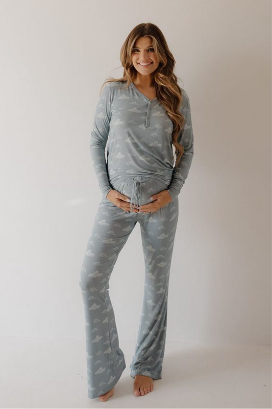 A smiling woman with long brown hair stands barefoot against a plain backdrop, wearing Kendy 3 Women's Bamboo Pajama | Duke in light blue with a cloud pattern. One hand is on her belly, the other at her side.