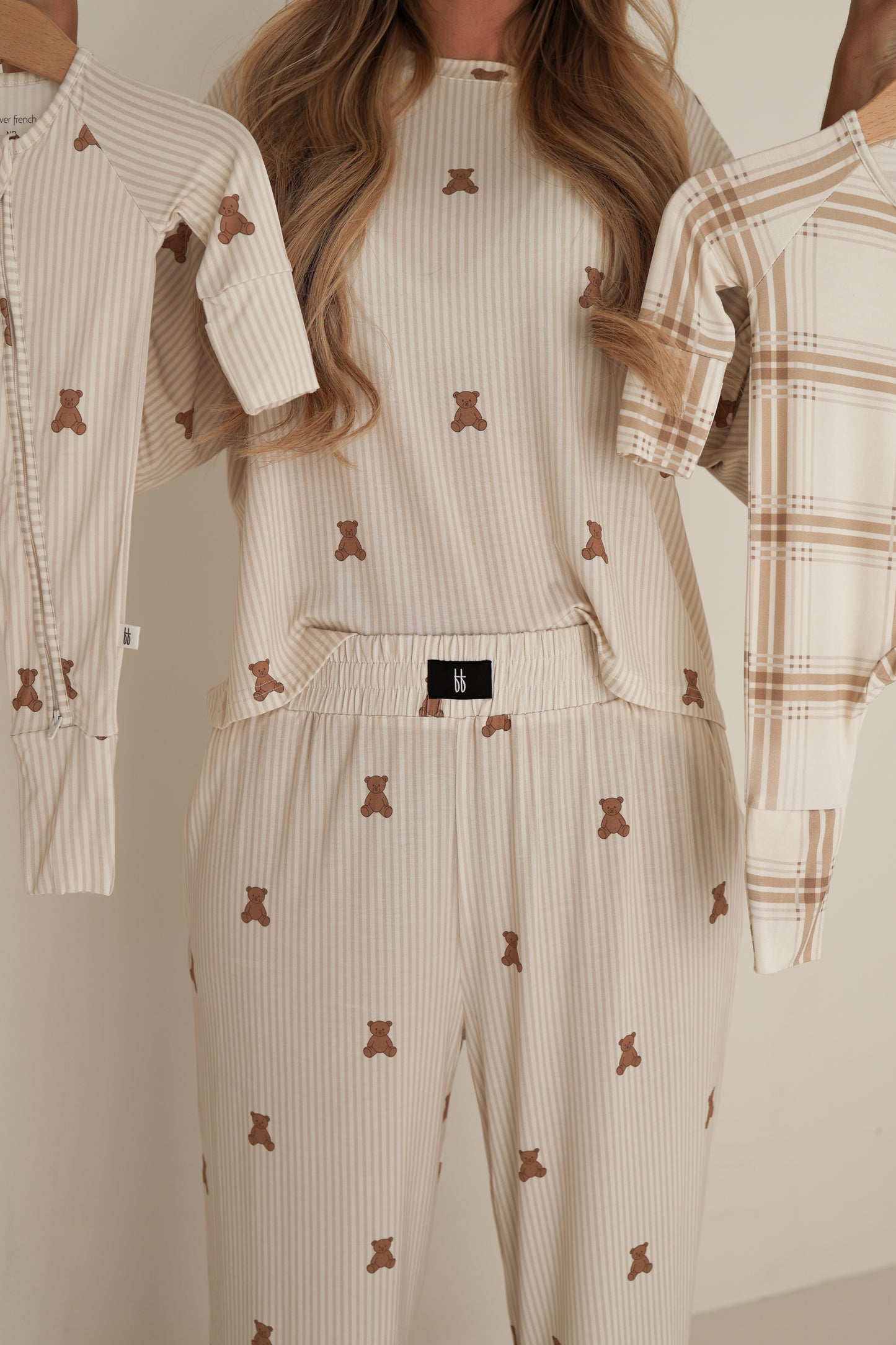 Wearing the forever french baby Women's Boxy Tee Lounge Set | Teddy, a person holds up two matching children's pajamas, one in each hand. Their cream-colored bamboo set features brown teddy bear prints. The person's face is not visible.