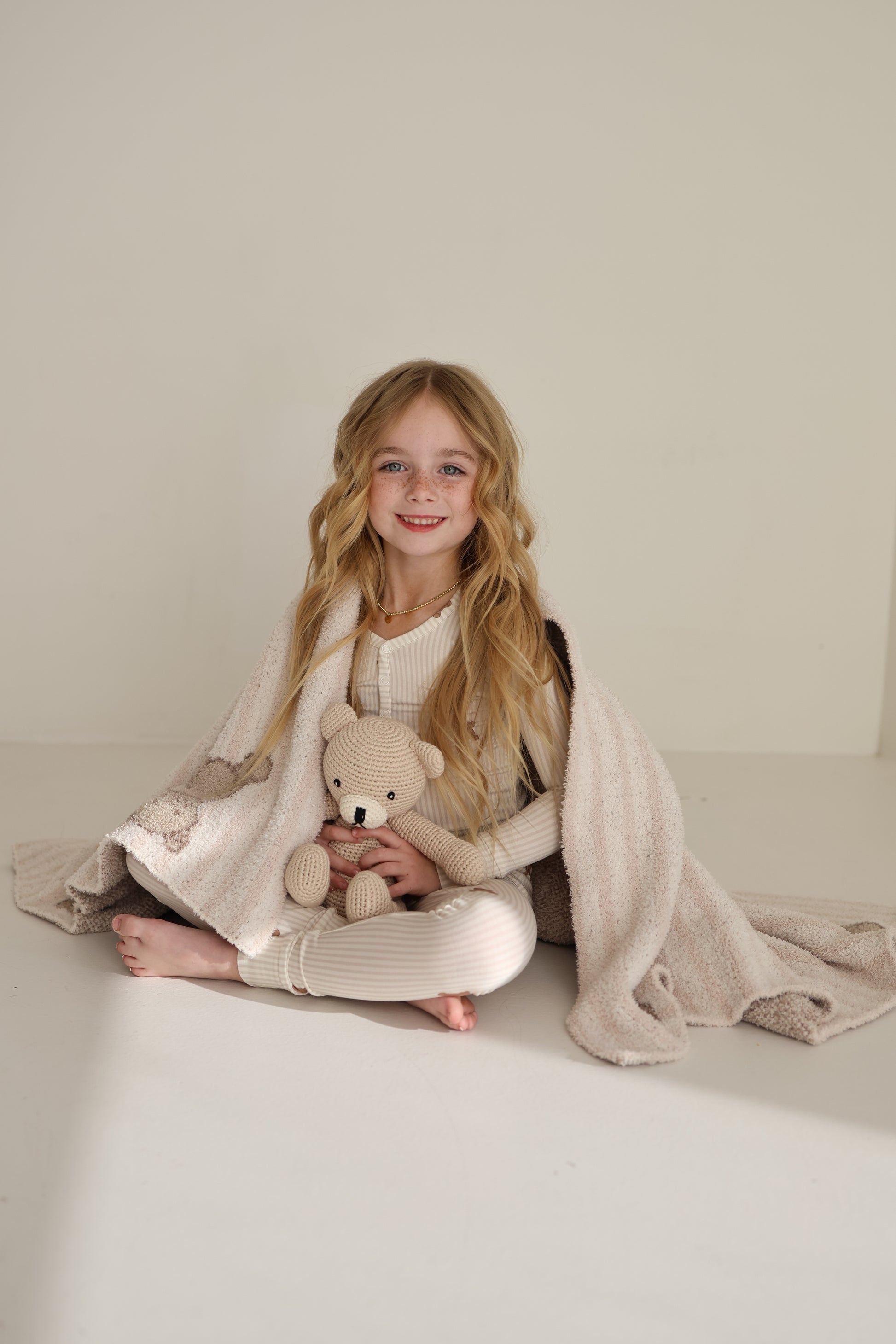 A young girl with long blonde hair sits barefoot on the floor, wrapped in a forever french baby Plush Blanket | Teddy, holding a knitted teddy bear and smiling at the camera in a bright, minimalist room.