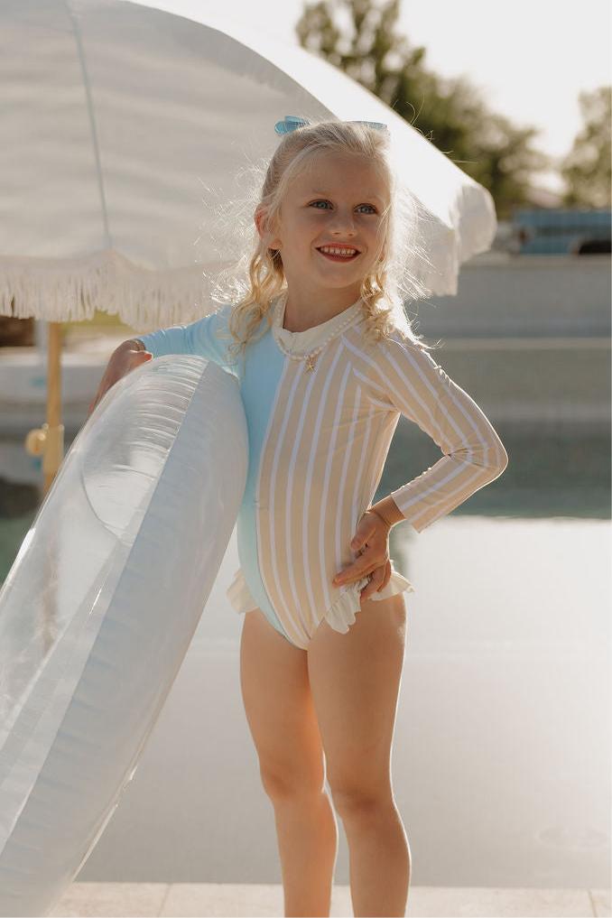 A young girl in a lolo webb 2 Girl Long Sleeve Swimsuit | Surf & Sand stands by a pool, smiling and holding a large clear inflatable ring, with a white fringed umbrella in the background.