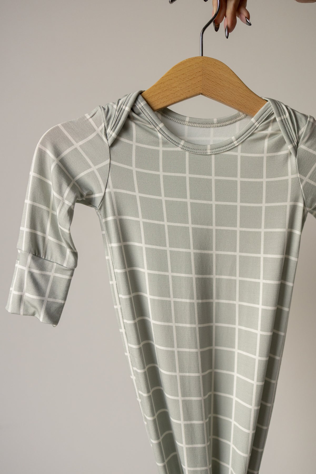 The Bamboo Knotted Gown | Foggy Grid from forever french baby, featuring a light gray color with a white grid pattern and made from hypoallergenic material, hangs on a wooden hanger held by a hand with manicured nails against a plain light background.