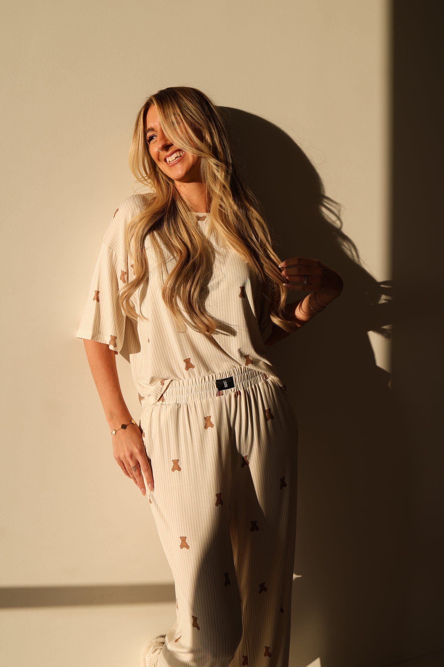 A woman with long, wavy blonde hair smiles against a light wall, wearing the forever french baby Women's Boxy Tee Lounge Set in Teddy. Sunlight casts soft shadows on the wall behind her.