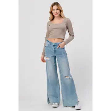 A person with long hair poses wearing a taupe long-sleeve crop top and light blue distressed High Rise Skater Jeans in Medium Wash from I&M JEAN, INC., paired with white sneakers. The background is plain white.