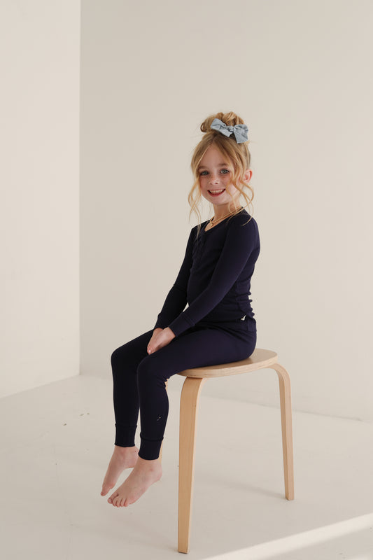 A young girl sits barefoot on a stool, smiling in forever french baby’s Bamboo Two Piece Pajamas | Blueberry Ribbed—hypoallergenic, ultra-comfy kidswear perfect for relaxing in a bright, minimalist room.