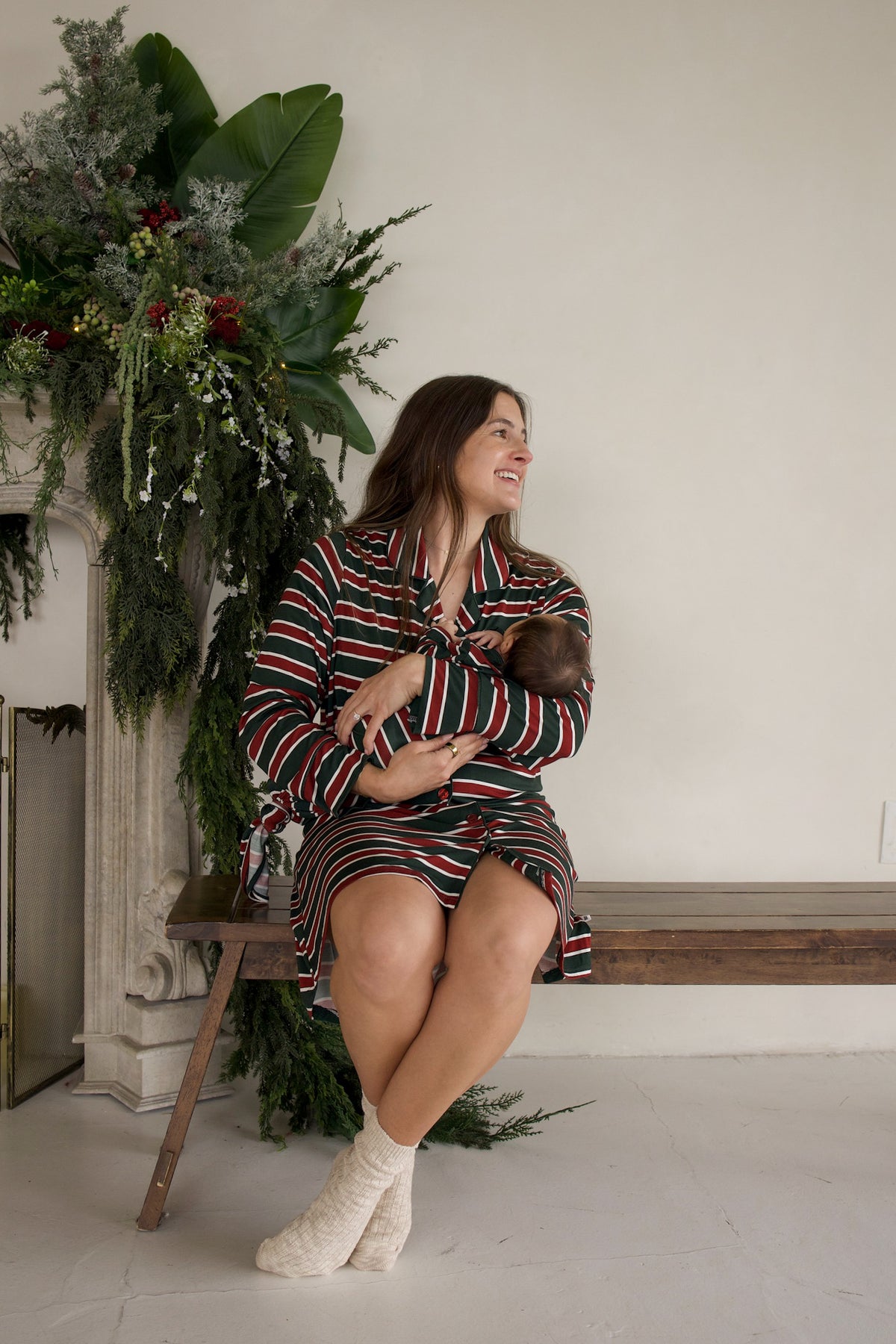 A woman wears a Women’s Bamboo Sleeping Dress | Holiday Stripe in red, green, and white while sitting on a wooden bench and cradling a newborn infant.