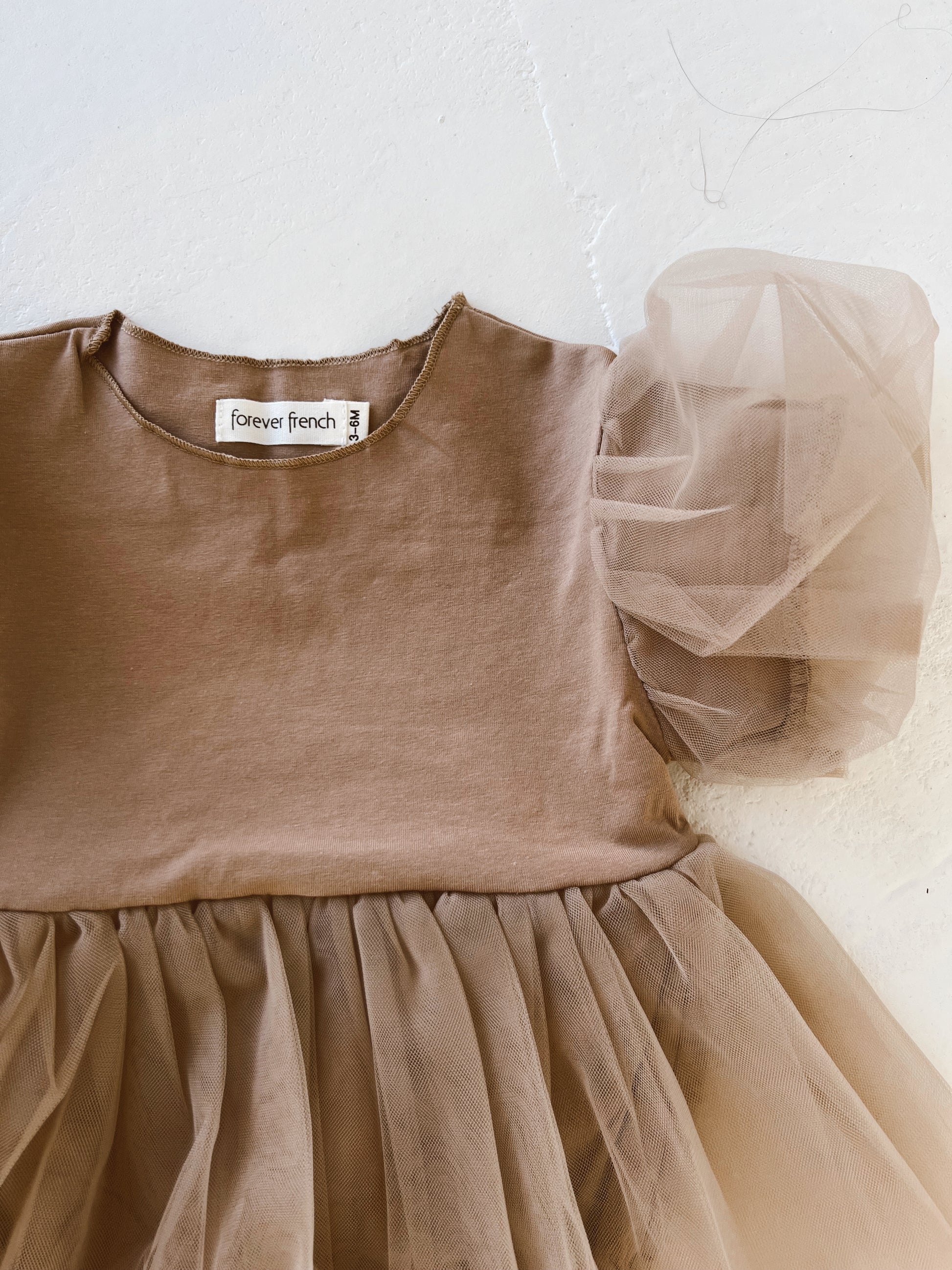 A close-up photo of a Taupe | Puff Sleeve Snap Suit Dress for children from forever french baby. The top is made of plain fabric, while the bottom features a voluminous tulle skirt. One side showcases a puff sleeve design in tulle, with a label inside the neckline reading "forever french baby".