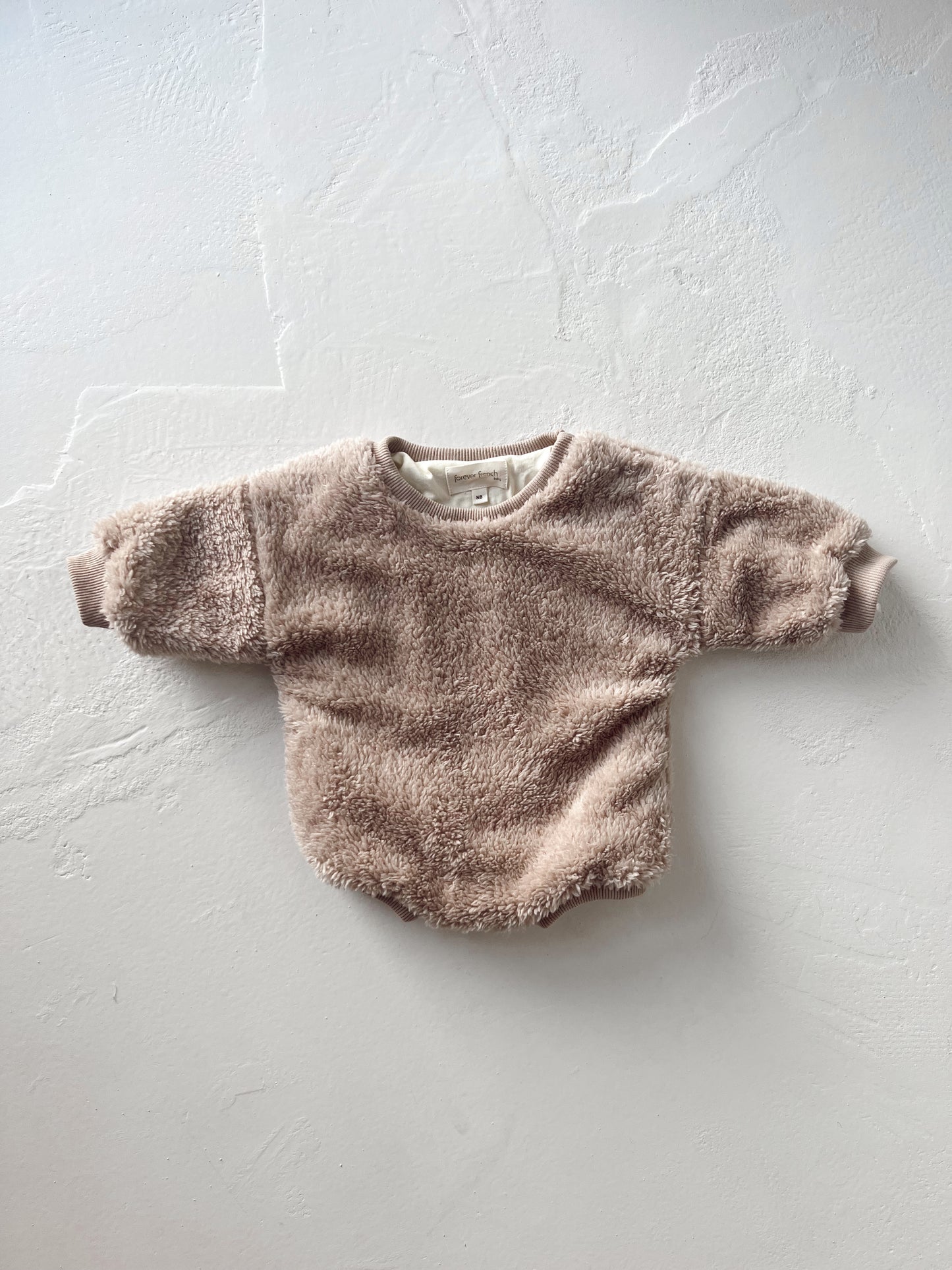 A cozy Long Sleeve Romper | Teddy Bear from forever french baby, featuring a light brown hue and fuzzy material with long sleeves, is laid flat against a white textured background. The baby romper includes ribbed cuffs and a crew neckline.