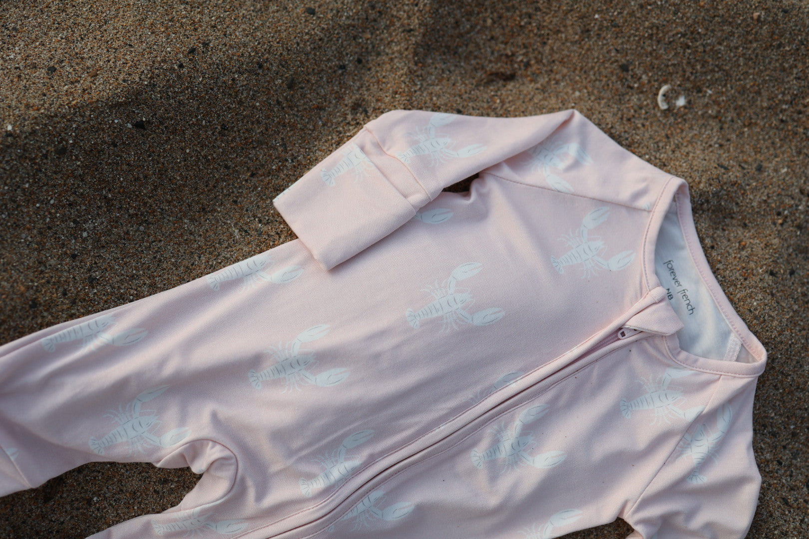 A pale pink Bamboo Zip Pajamas | Lobster by forever french baby, made from hypoallergenic fabric and featuring a white lobster print, lies partially buried on sandy ground.