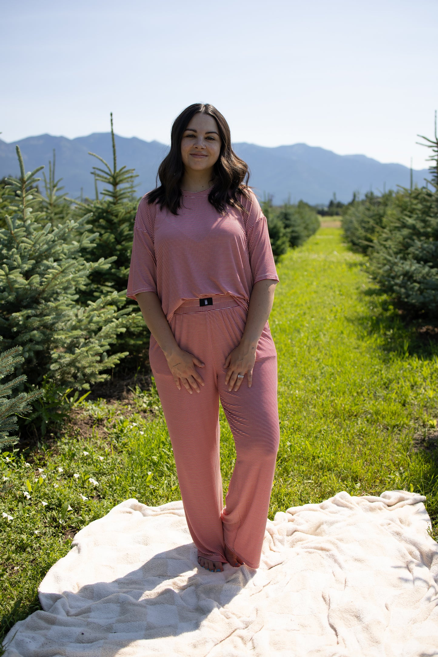 A woman wearing the forever french baby Women's Boxy Tee Lounge Set in Peppermint Stripe Ribbed stands barefoot on a blanket outdoors, surrounded by small pine trees and mountains in the background under a clear sky.