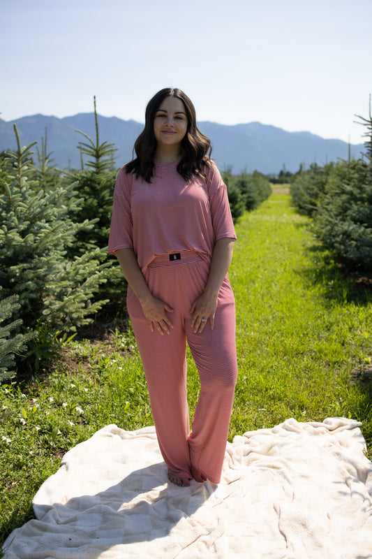 A woman wearing the forever french baby Women's Boxy Tee Lounge Set in Peppermint Stripe Ribbed stands barefoot on a blanket outdoors, surrounded by small pine trees and mountains in the background under a clear sky.