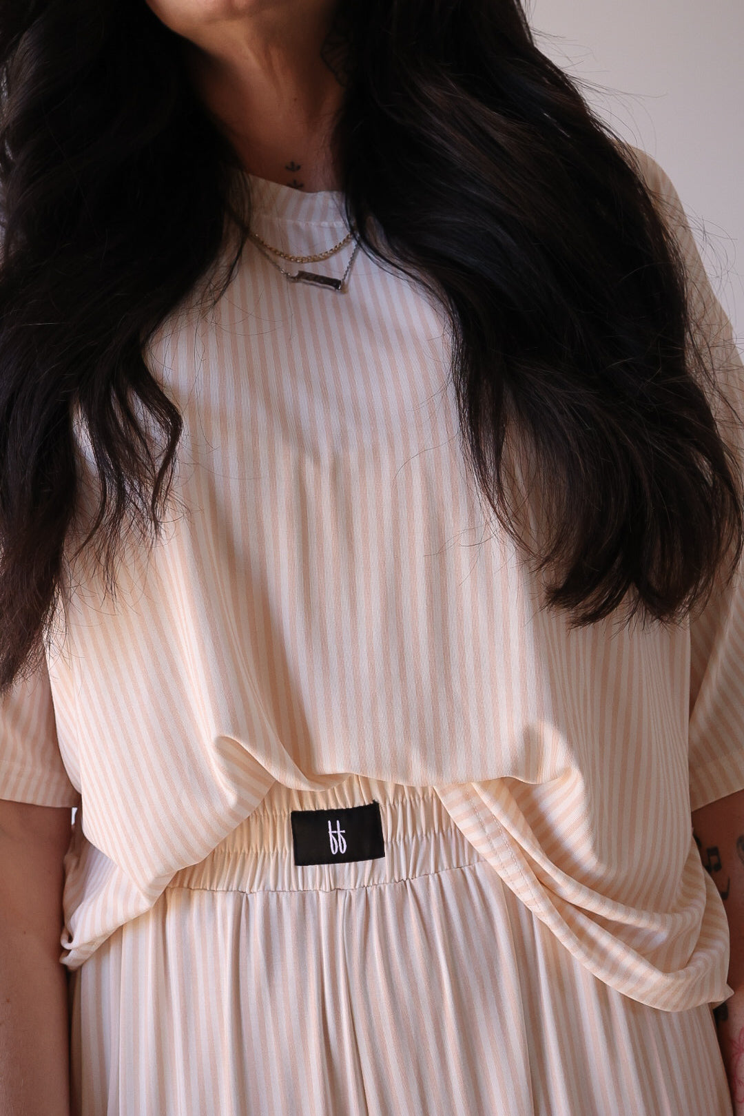 A woman with long dark hair wears the Women's Boxy Tee Lounge Set in Sadie Stripe by forever french baby, featuring a light-colored, short-sleeve, vertically striped top tucked into a matching skirt with a visible "bb" black waistband label.