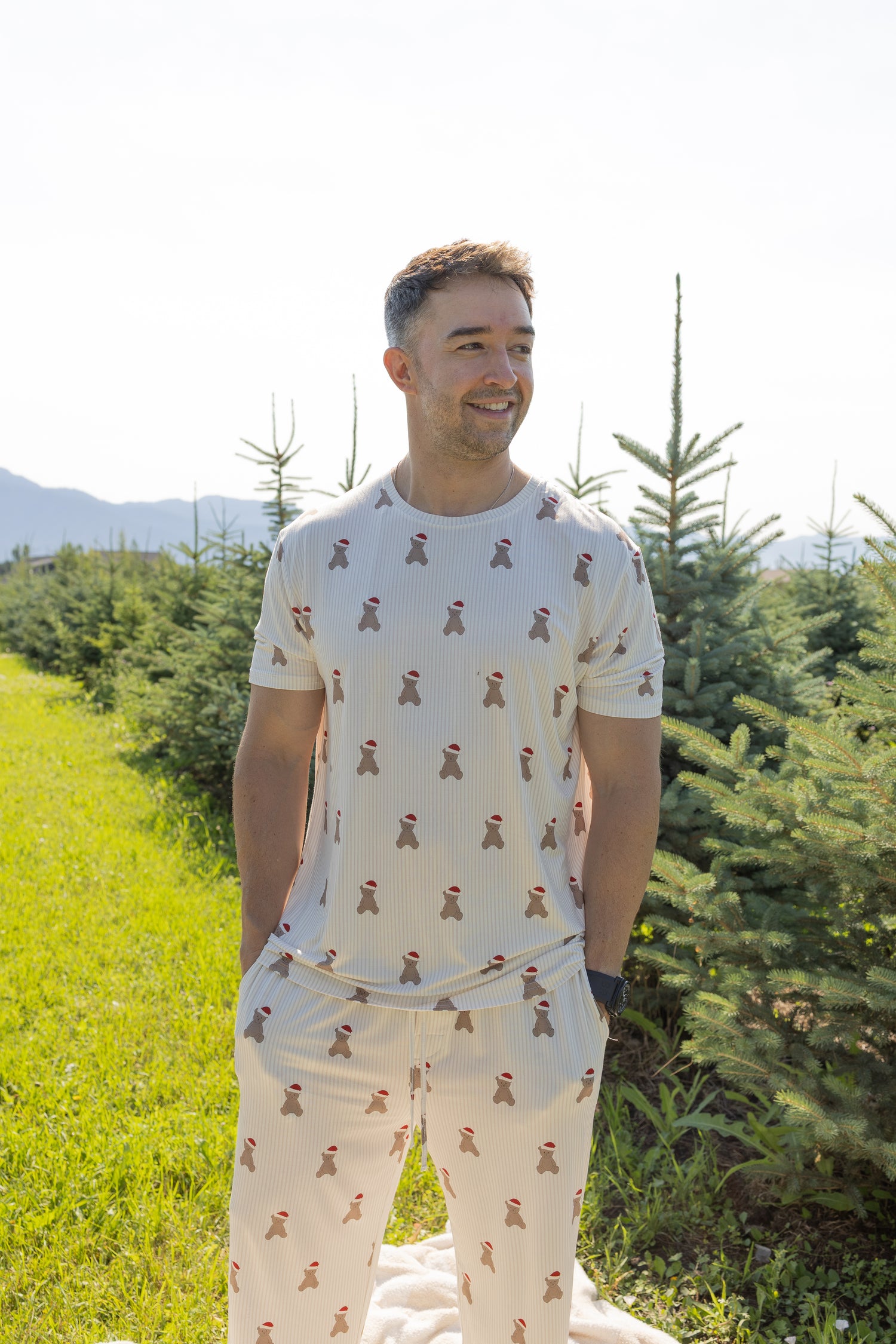 A man wearing the Men's Short Sleeve Pajamas in the Santa Teddy print from forever french baby stands in a sunny field beside pine trees, smiling and looking to the side.