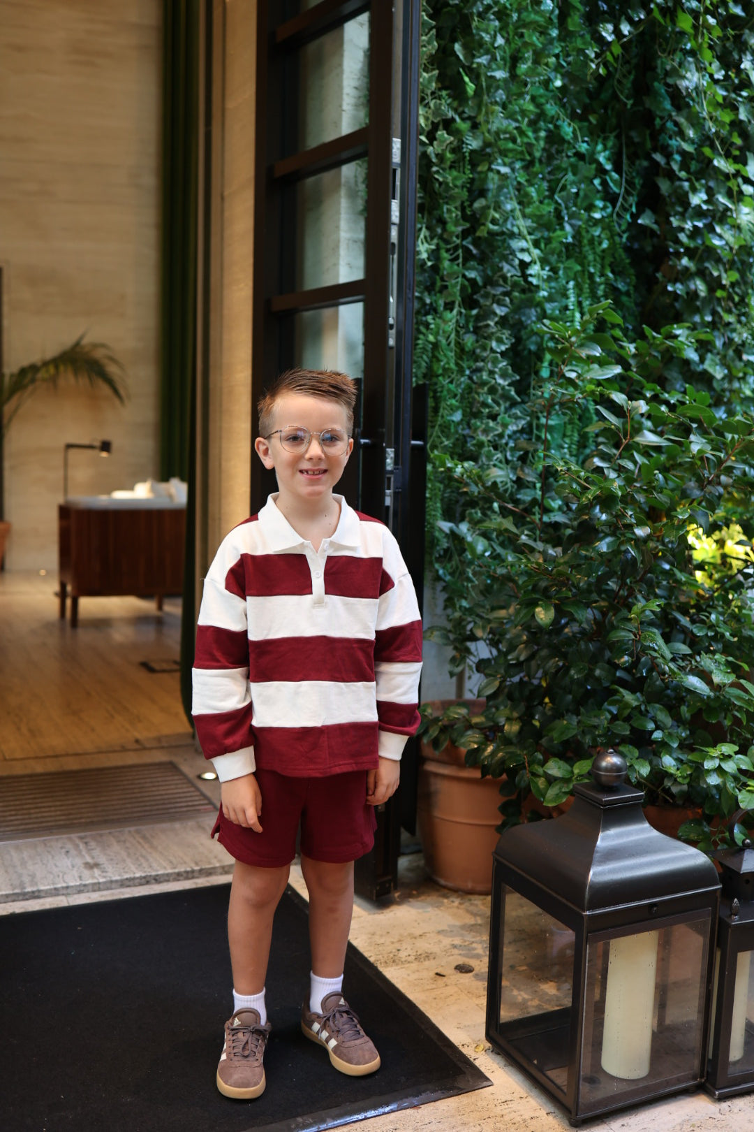 A young boy wearing a white and cranberry-striped long-sleeve rugby shirt and matching Child Terry Shorts | Deep Cranberry.