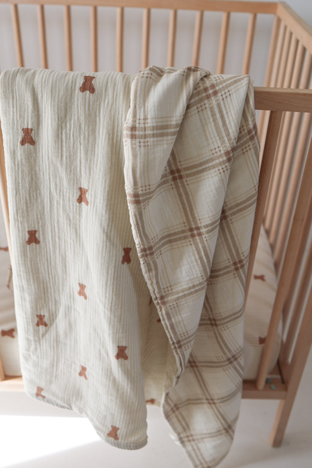 Two forever french baby Muslin Quilt | Teddy & Ellis Plaid bedding pieces hang over a light wood crib: one with a teddy bear print, the other in beige and brown plaid, plus a striped crib sheet inside.