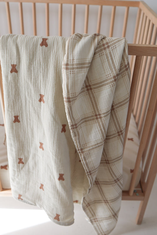 Two forever french baby Muslin Quilt | Teddy & Ellis Plaid bedding pieces hang over a light wood crib: one with a teddy bear print, the other in beige and brown plaid, plus a striped crib sheet inside.