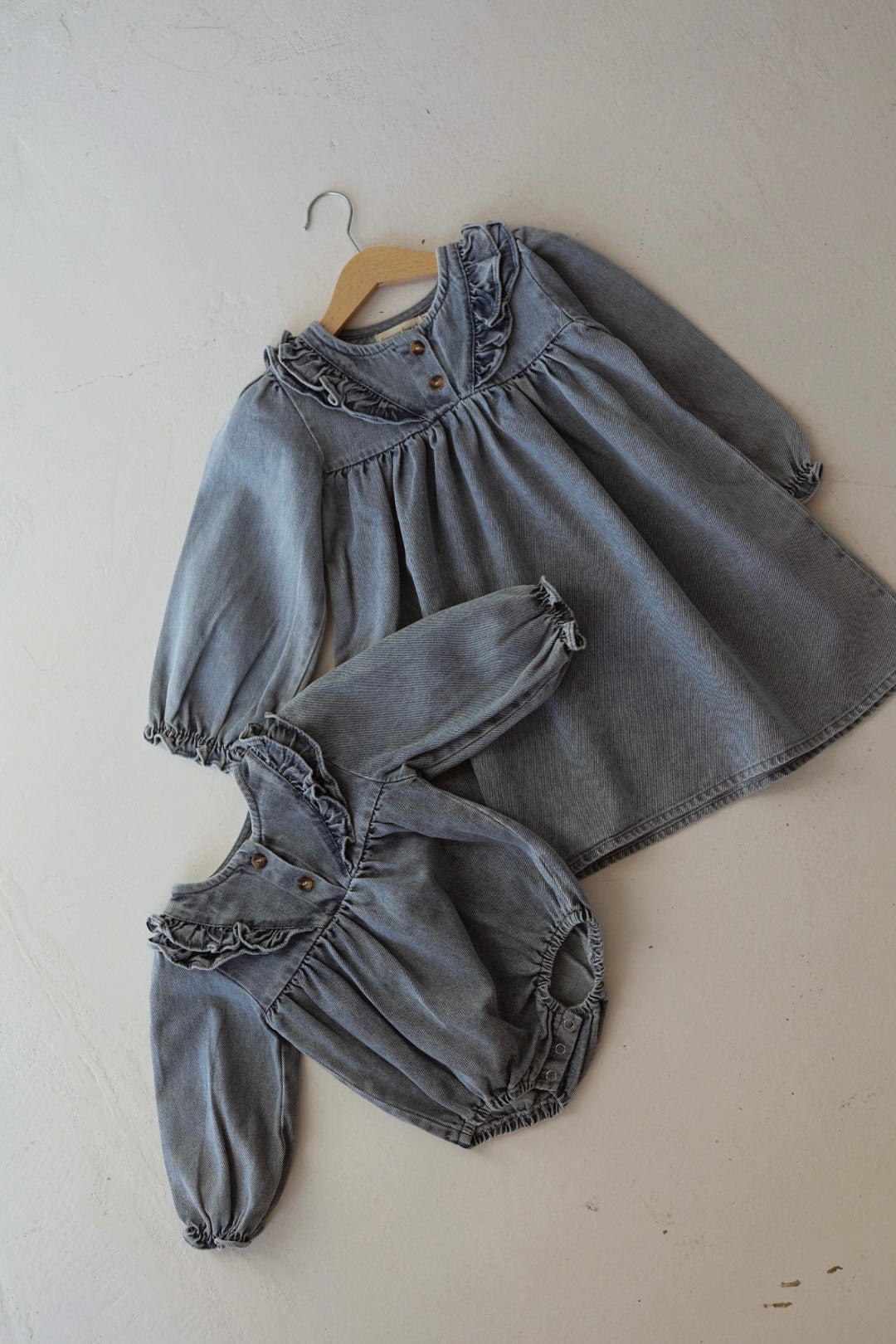 Two matching gray denim children's outfits from forever french baby lie flat: the Denim Dress | Ruffle, a tunic-style long sleeve dress with ruffles and wooden buttons, and a similar romper. Both have gathered cuffs and hems for a coordinated look.