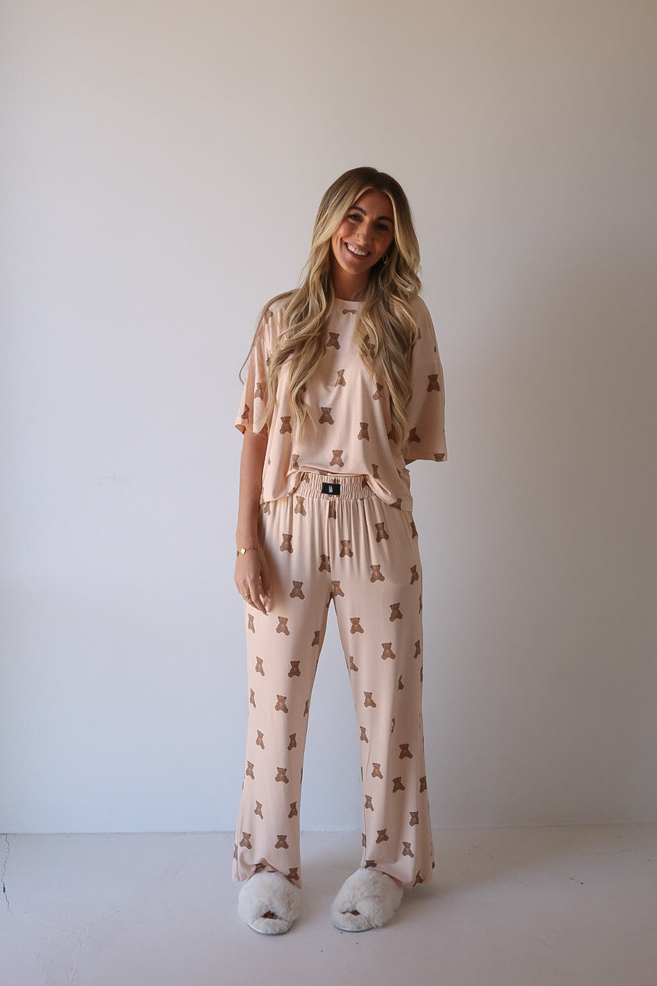 A smiling woman with long blonde hair wears the forever french baby Women's Boxy Tee Lounge Set | Lottie in light pink with a brown bear print and fluffy white slippers, standing against a plain, light-colored wall.