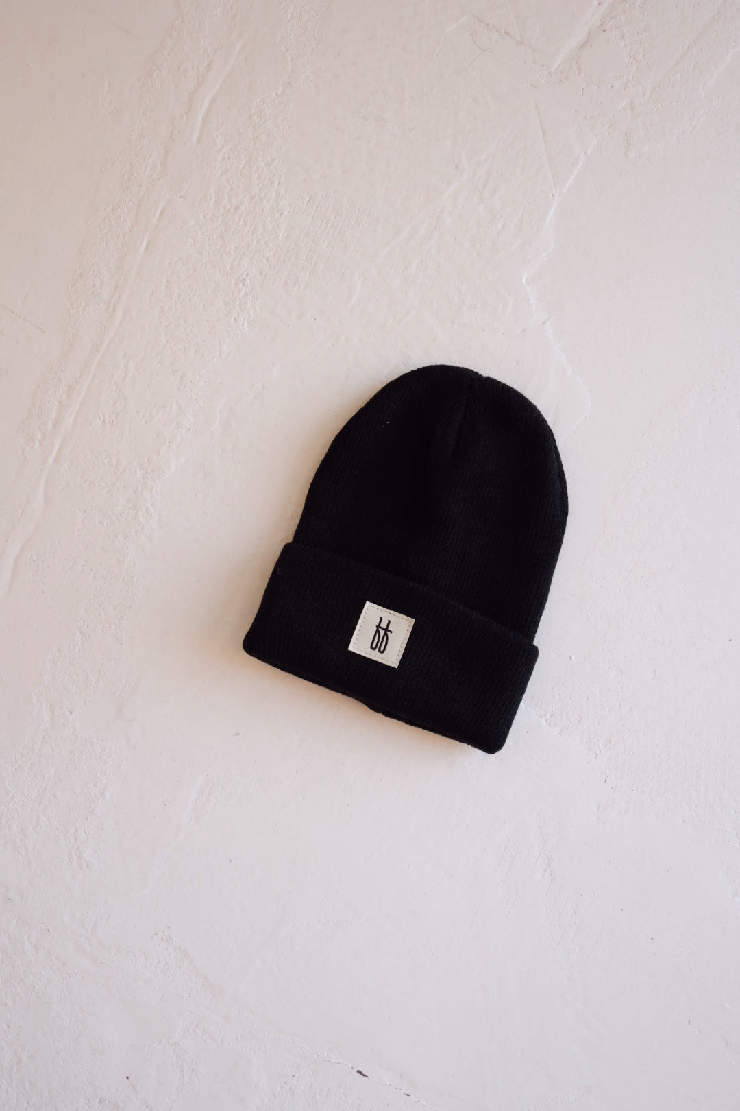 A black ff Knit Beanie from forever french baby with a folded brim rests on a textured off-white surface. The beanie features a small rectangular tag with a minimalist design near the edge of the folded brim and boasts a subtle ribbed design.