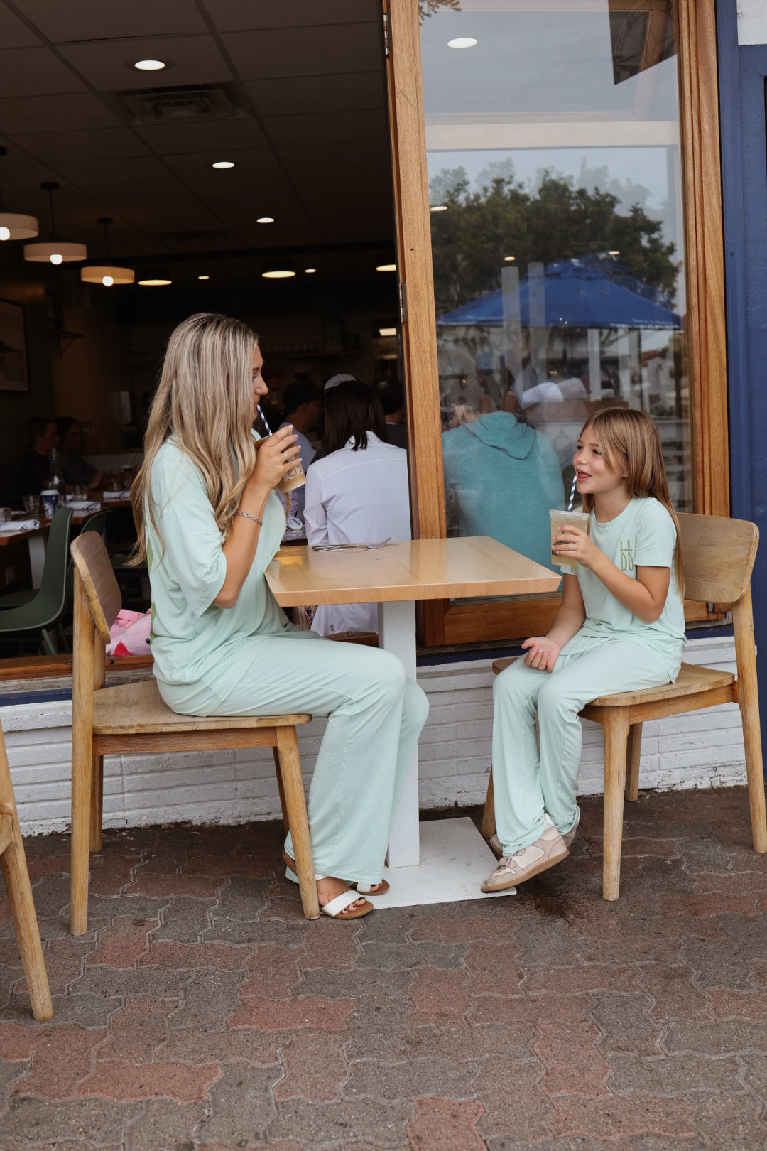 A smiling woman and young girl enjoy iced drinks at an outdoor café, both dressed in comfortable, matching forever french baby Meet Me at the Beach bamboo lounge sets, perfect for relaxing together in style.