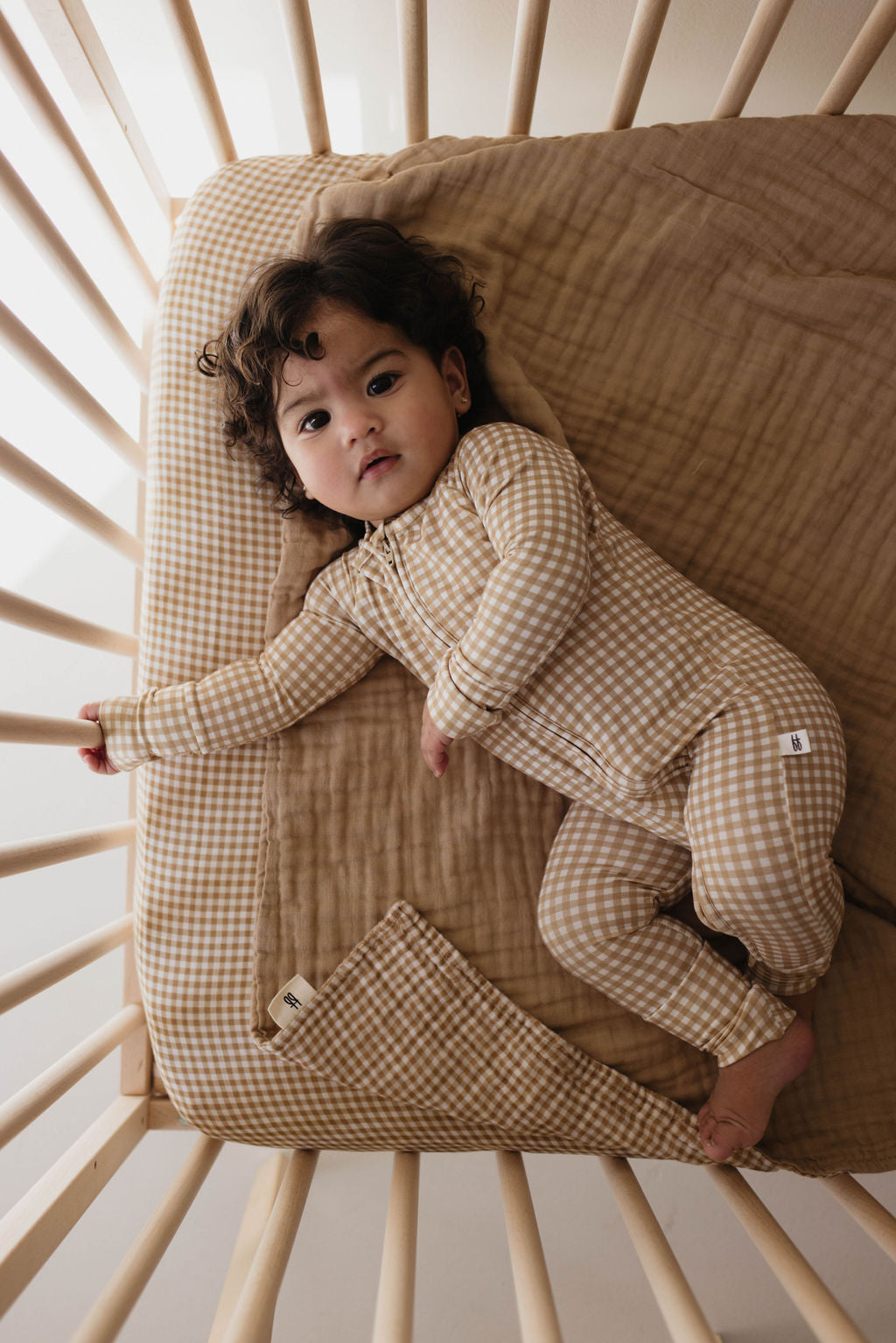 A curly-haired baby lies in a crib, wearing forever French baby's Bamboo Zip Pajamas in Golden Gingham, crafted from breathable fabric. The crib sports a matching checkered sheet and a brown blanket. The baby is comforted by hypo-allergenic clothing with one arm extended upward.