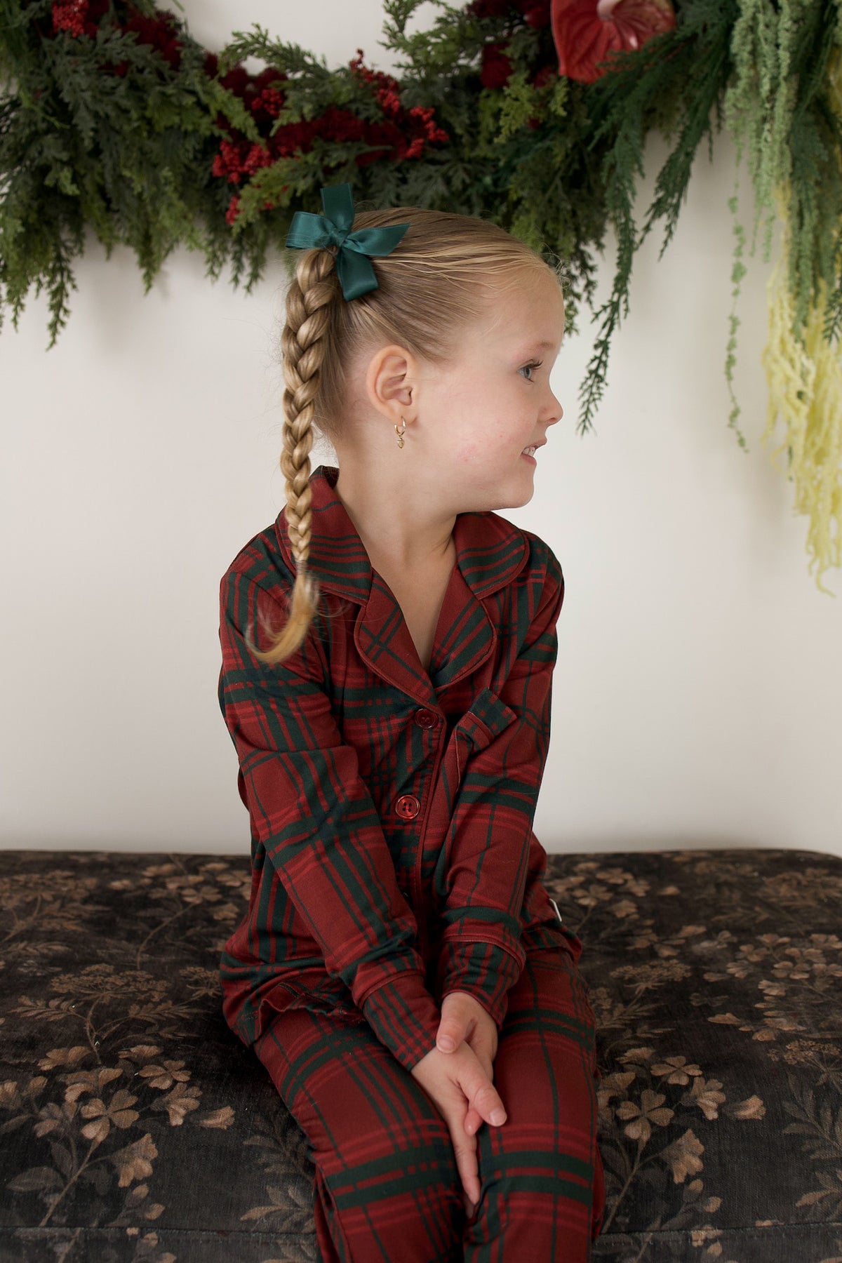 A young girl wears the Girl’s Bamboo Button Up Lounge Set | Christmas Plaid by forever french baby, sitting sideways on a bench with a green bow in her braid. A festive green and red holiday wreath hangs behind her.