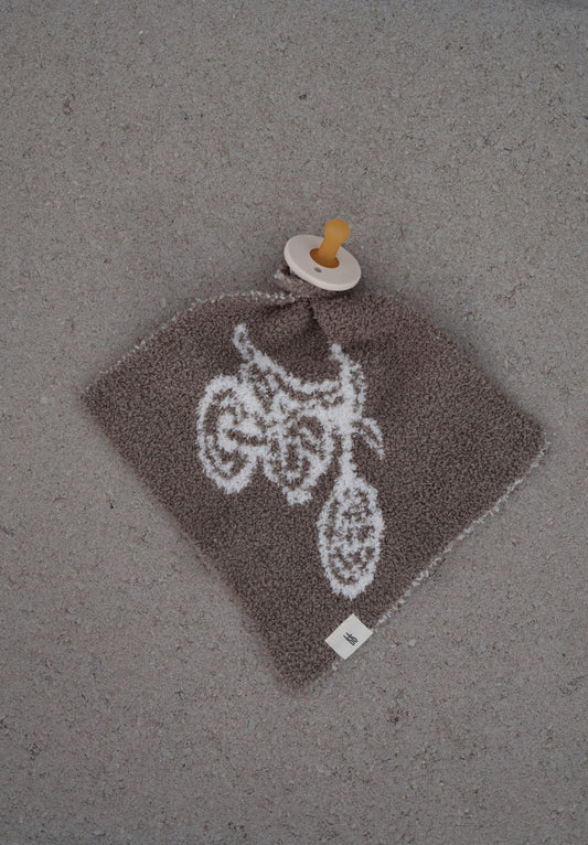 A Lovey | Dirt Bike by forever french baby, featuring a brown textured fabric with a white dirt bike design and attached to a yellow pacifier, rests on a gray surface.