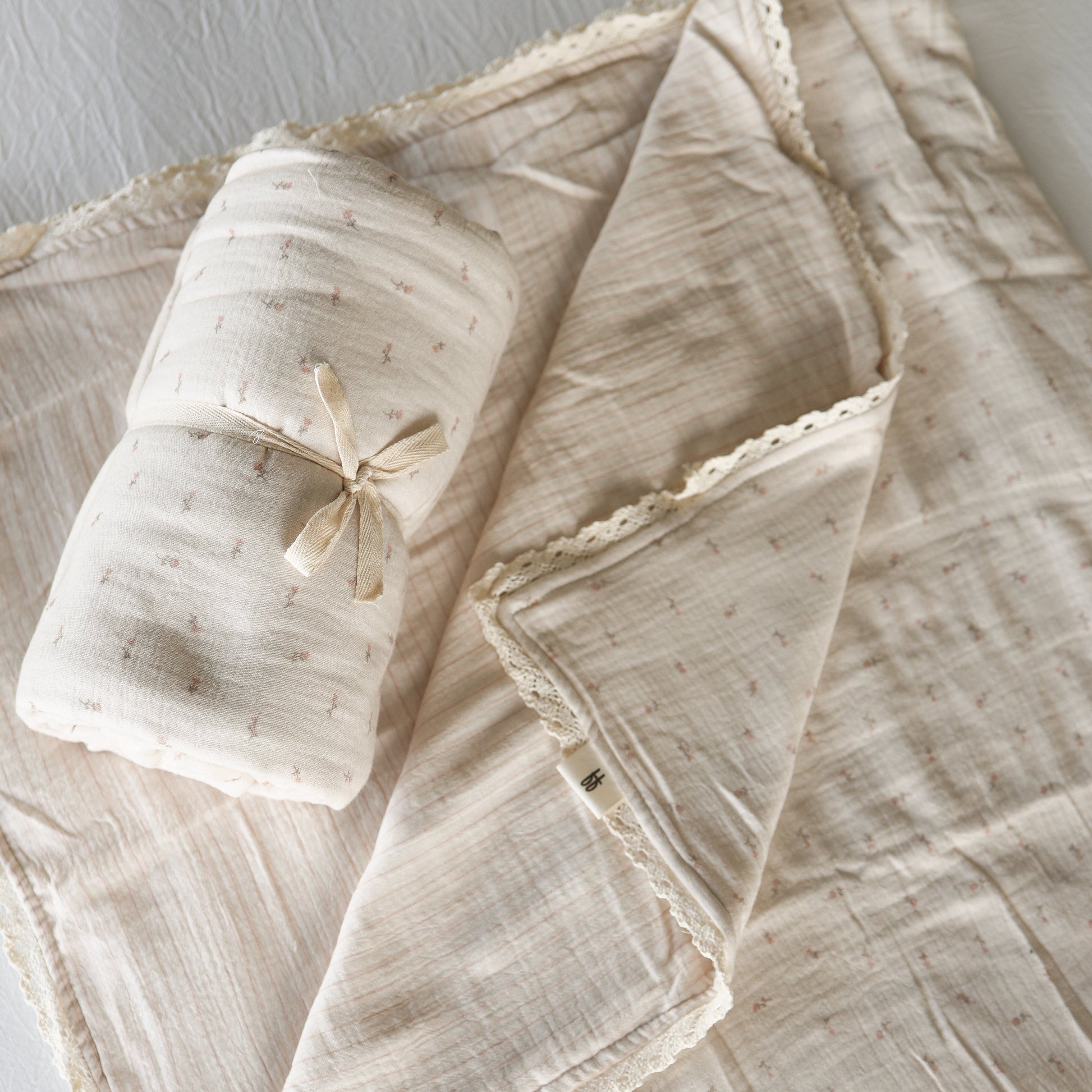 The forever french baby Muslin Quilt | Lucy, in soft cream 100% cotton with lace trim and a subtle dotted pattern, is shown flat beside a matching rolled-up quilt tied with ribbon—perfect for nursery bedding.