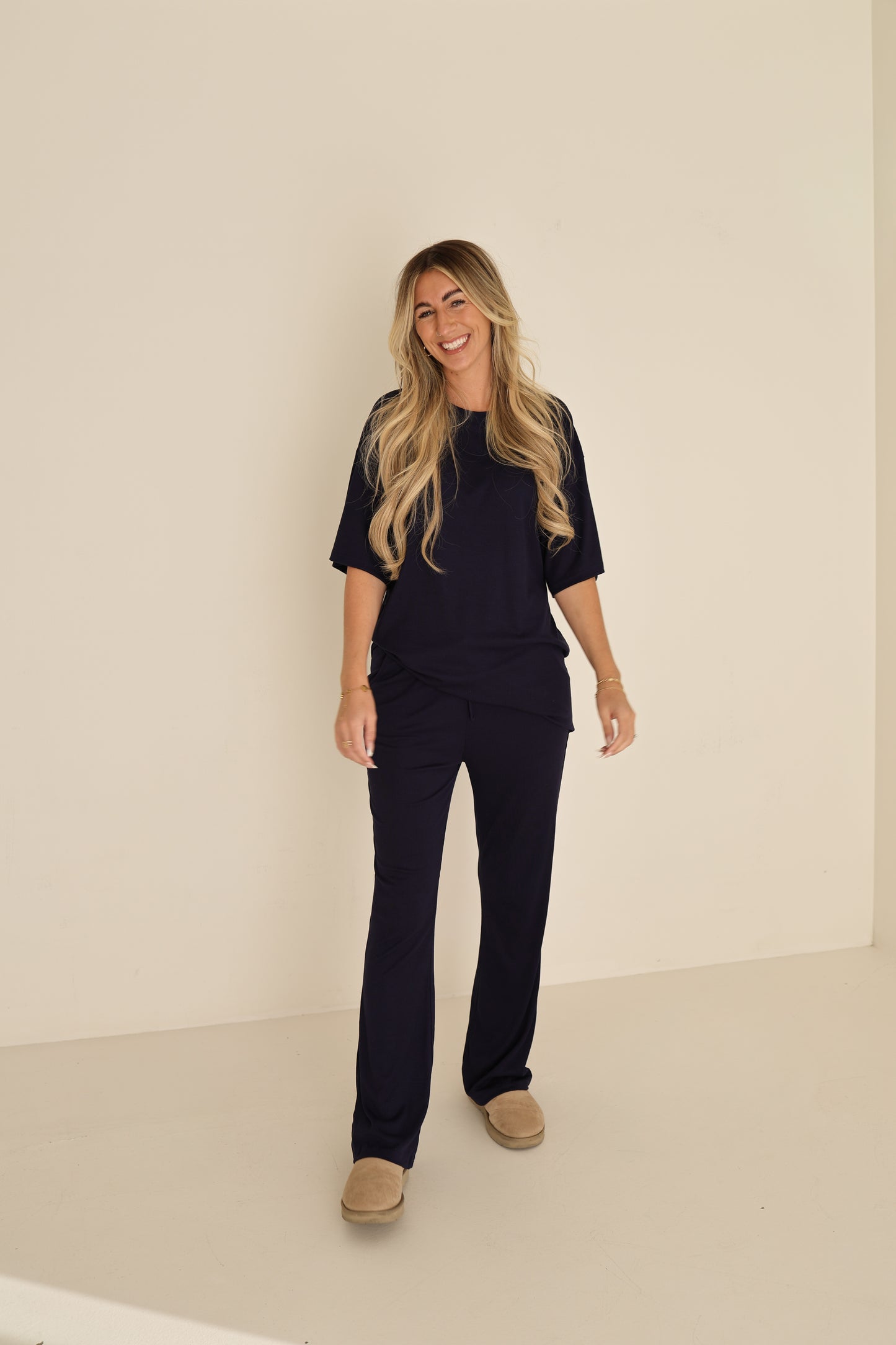 A woman with long blonde hair smiles against a plain light wall wearing the forever french baby Short Sleeve Bamboo Women's Lounge Set in Blueberry Ribbed with beige slippers.