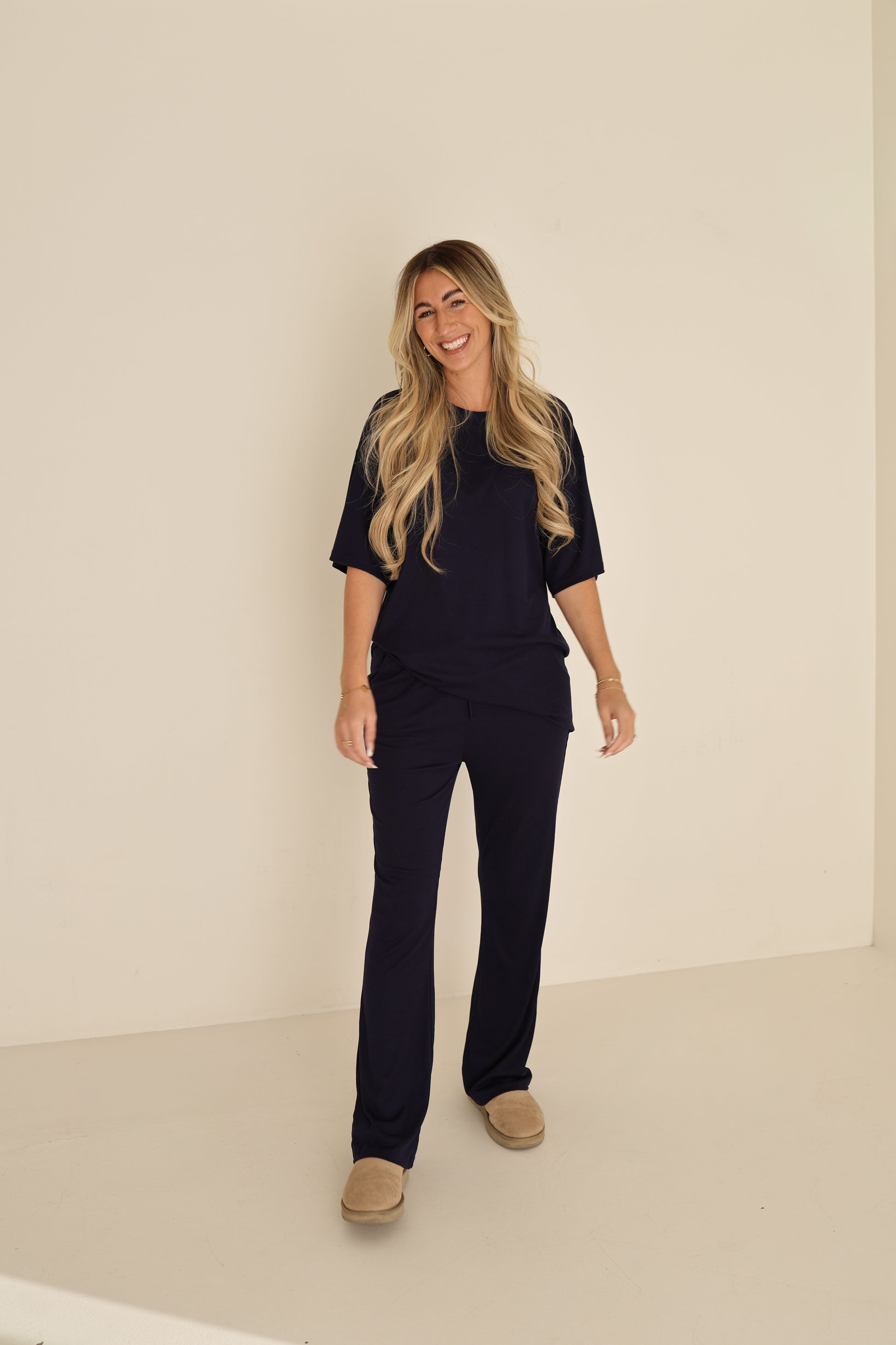 A woman with long blonde hair smiles against a plain light wall wearing the forever french baby Short Sleeve Bamboo Women's Lounge Set in Blueberry Ribbed with beige slippers.