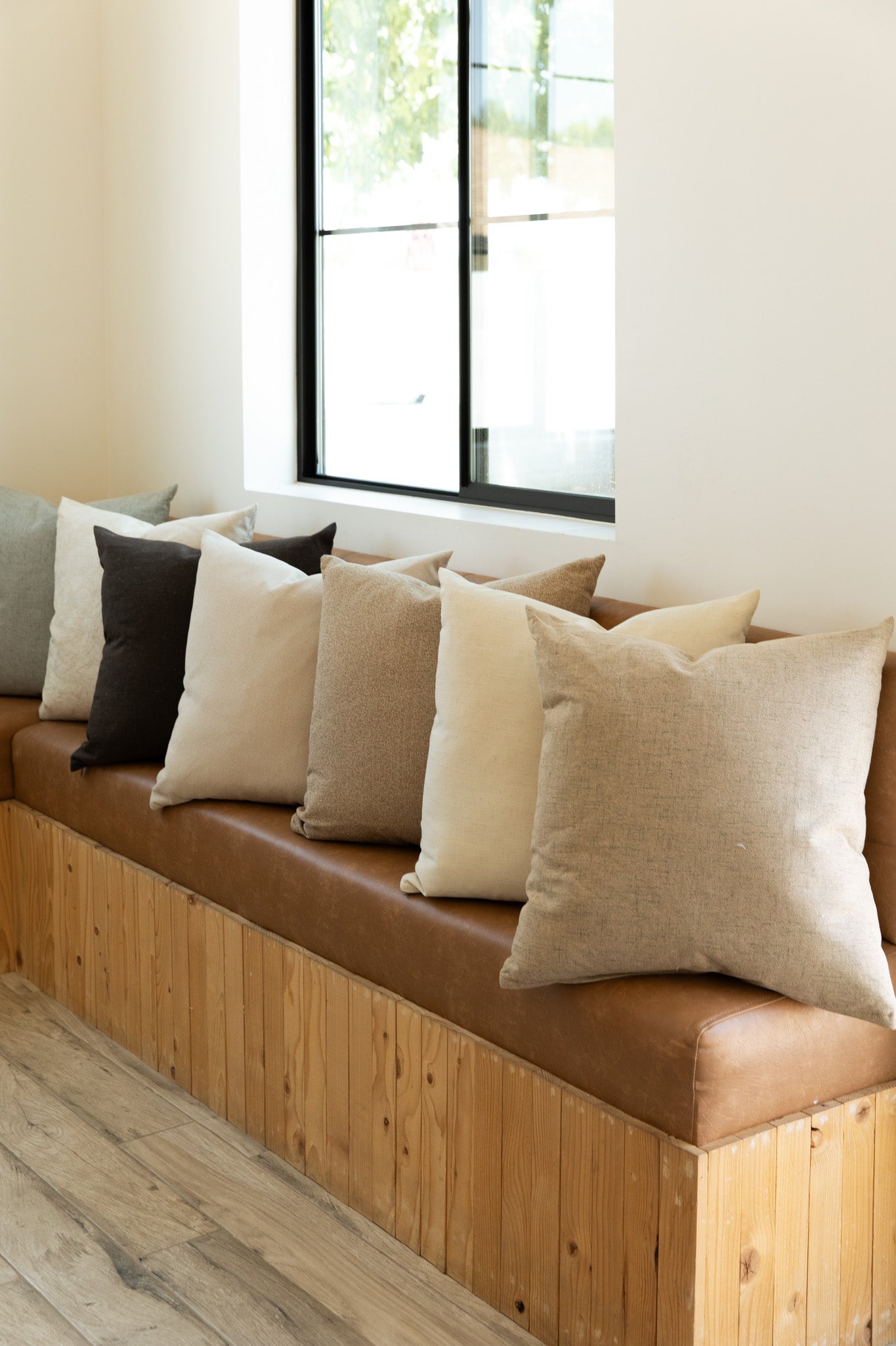 A row of Hazelnut | French Pillow from forever french home, featuring beige, brown, and gray throw pillows with feather down inserts, is neatly arranged on a long wooden bench with a light brown cushion. The bench is placed against a wall with a black-framed window from Forever French Home, allowing natural light to illuminate the cozy seating area.