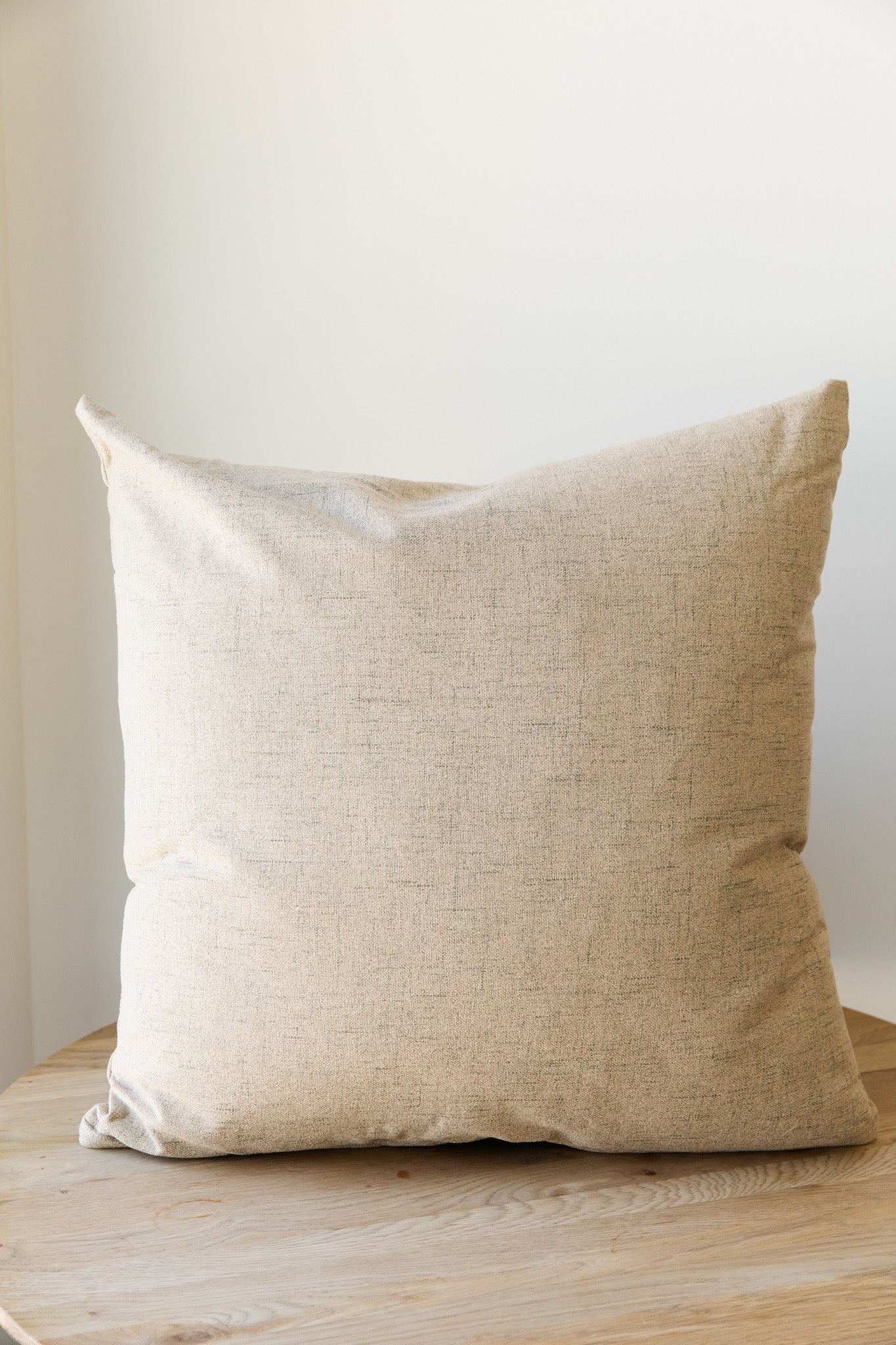 A beige square Hazelnut | French Pillow with a linen-like texture rests on a light wooden surface against a plain white wall. The pillow, featuring a feather down insert from Forever French Home, appears soft and slightly wrinkled, giving it a cozy and relaxed look.