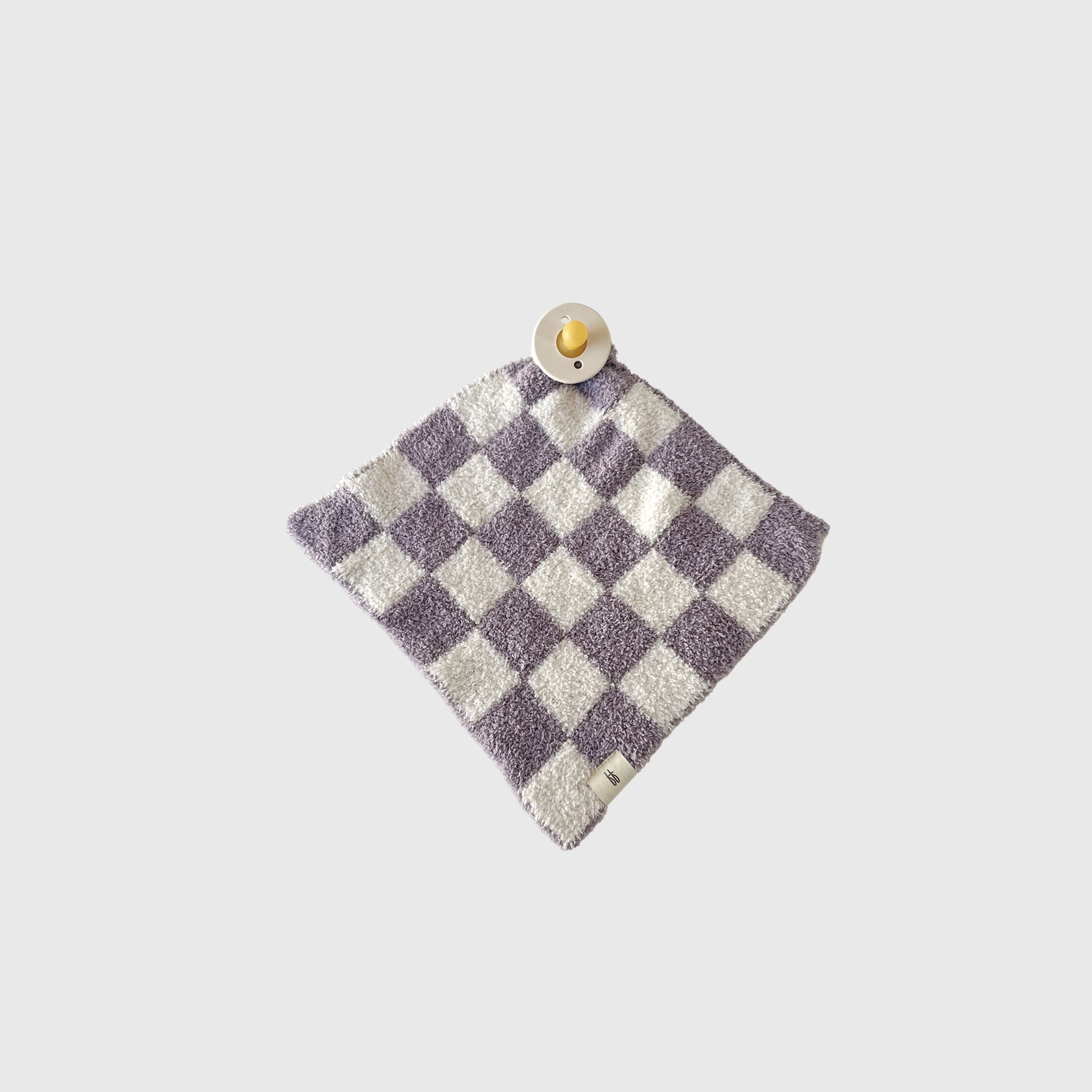 The Lovey | Grape Soda by forever french baby, a purple and white checkered microfiber blanket, is laid flat on a light gray background with a beige pacifier resting on one corner.