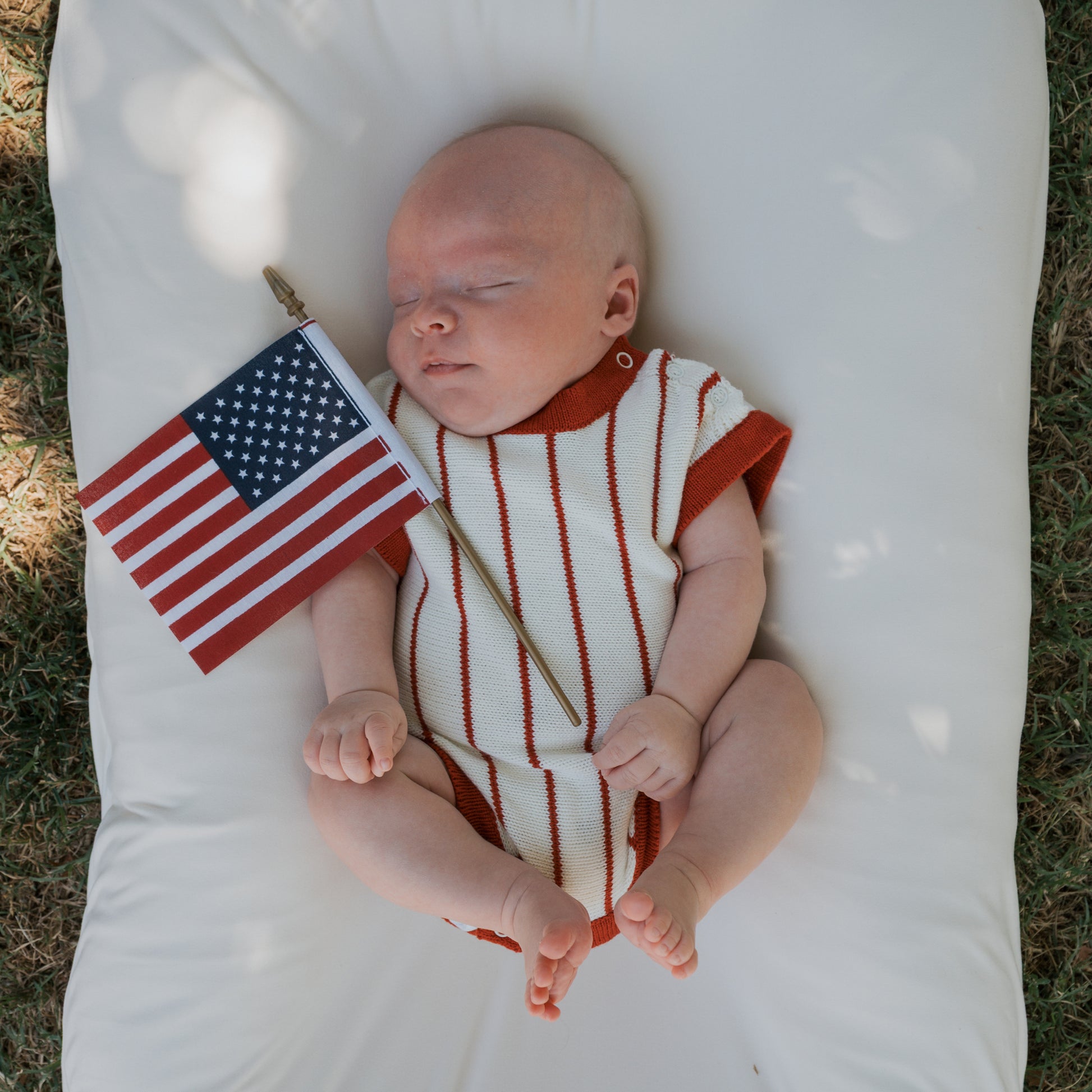 A baby wearing the forever french baby Knit Romper | Picnic Pinstripe with red and white stripes lies on a white cushion outdoors, holding a small American flag.
