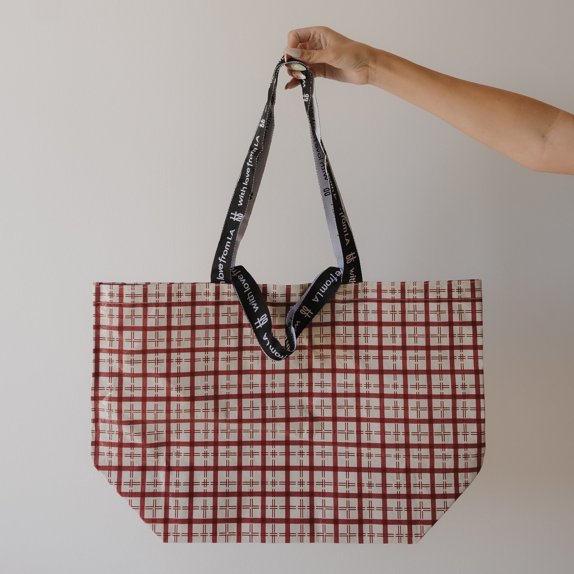 A hand holds up the ff Tote Bag | Window Pane by forever french baby, a large red and white checkered carry-all crafted from durable woven polypropylene with black handles, white text, shown against a plain white background.