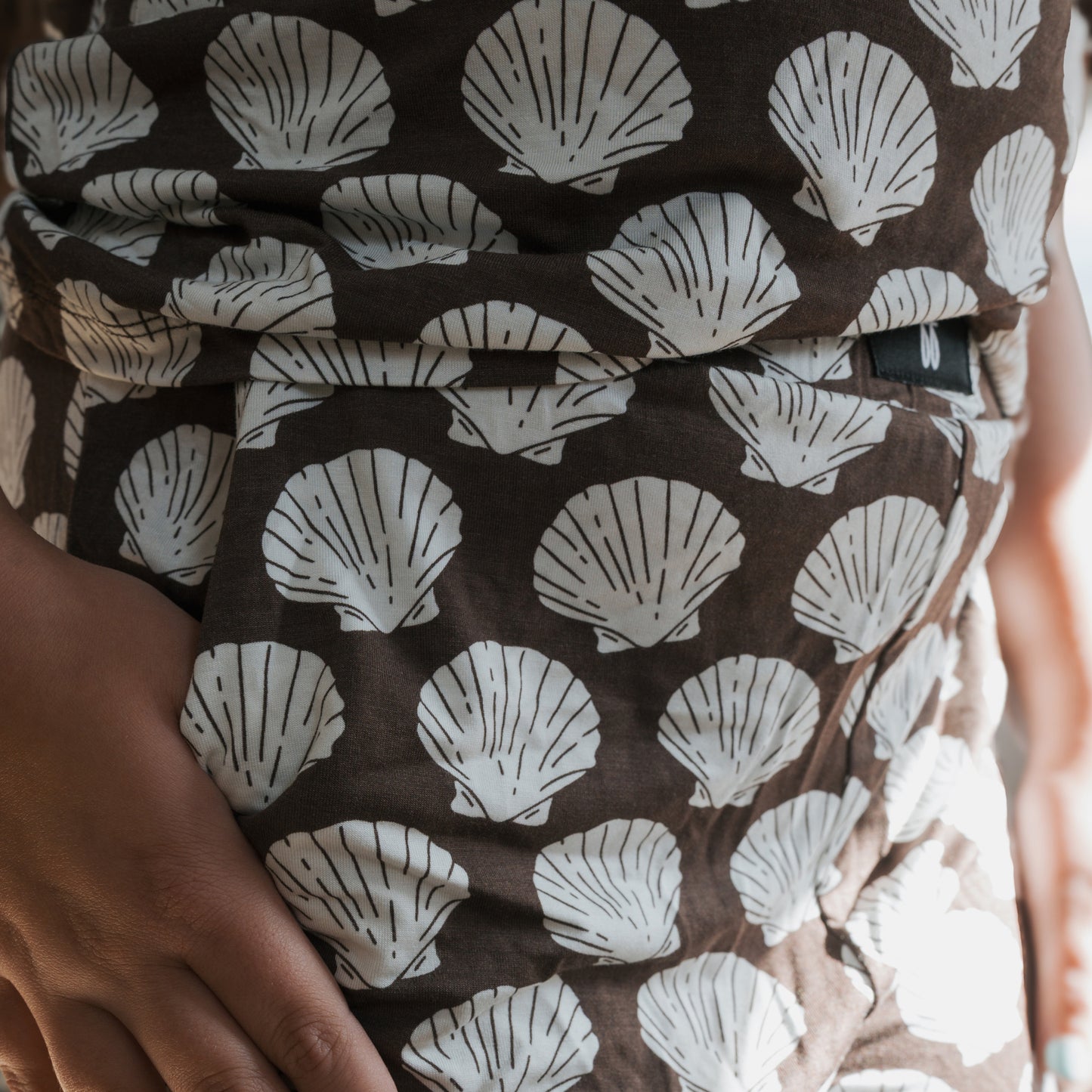 A close-up of a hand resting on a hip wearing the forever french baby Child Boxy Tee Lounge Set in brown, featuring breathable fabric with white seashell patterns highlighted by the light.