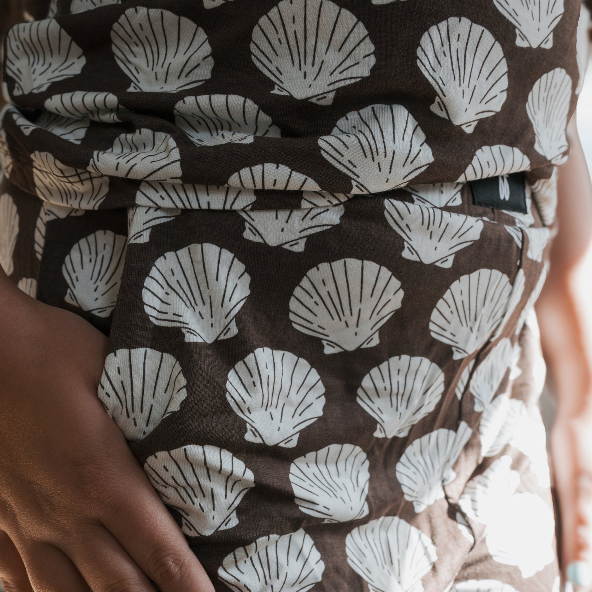A close-up of a hand resting on a hip wearing the forever french baby Child Boxy Tee Lounge Set in brown, featuring breathable fabric with white seashell patterns highlighted by the light.